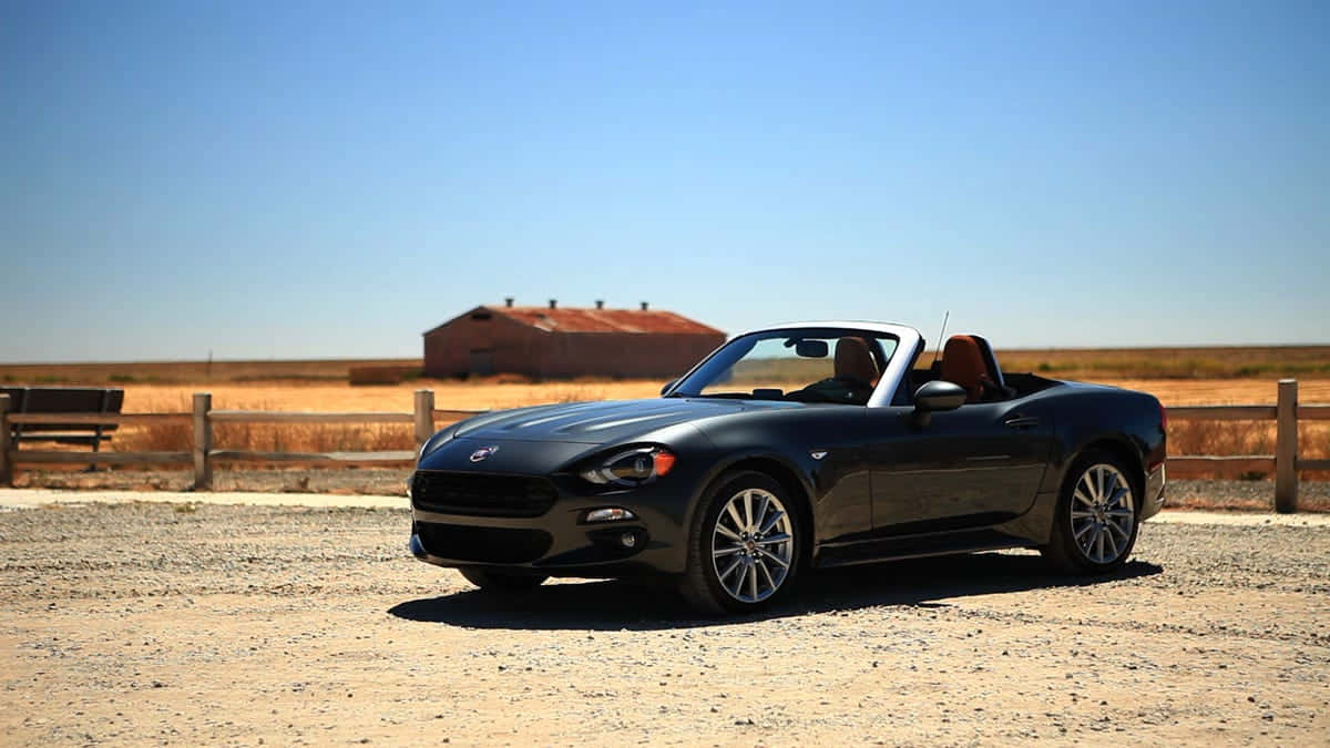 Fiat 124 Spider 桌布 壁紙 — 一架時尚黑色開篷跑車停泊在碎石地上，背景是晴朗藍天和一座質樸的紅色穀倉。