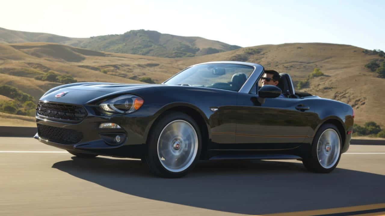 Fiat 124 Spider 桌布 壁紙 — 一架時尚的黑色Fiat 124 Spider開篷車在陽光明媚的高速公路上巡航，背景是連綿的金色山丘。