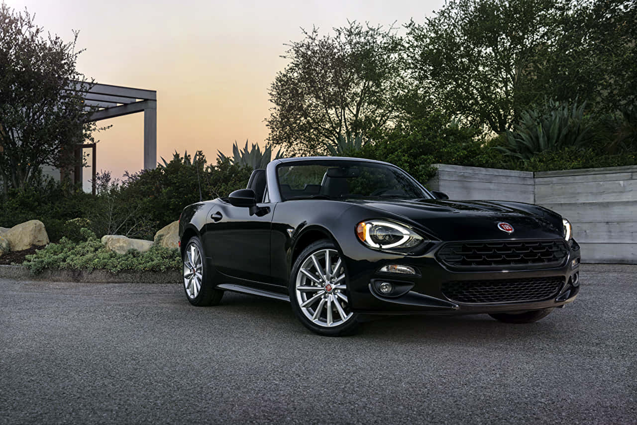 Fiat 124 Spider 桌布 壁紙 — 一架時尚的黑色Fiat 124 Spider開篷車停泊在鋪砌的車道上，背景是日落、樹木和一個現代化的涼棚。
