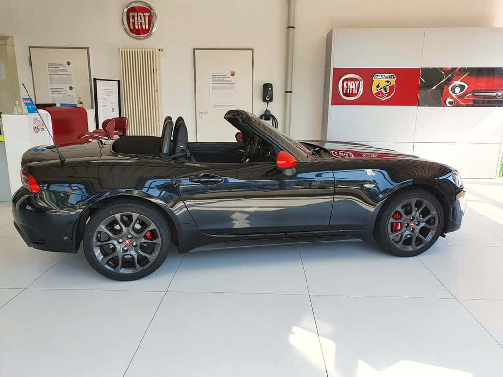 Fiat 124 Spider 桌布 壁紙 — 一輛時尚的黑色Fiat 124 Spider開篷車帶有紅色點綴，在明亮的經銷商陳列室中展示。