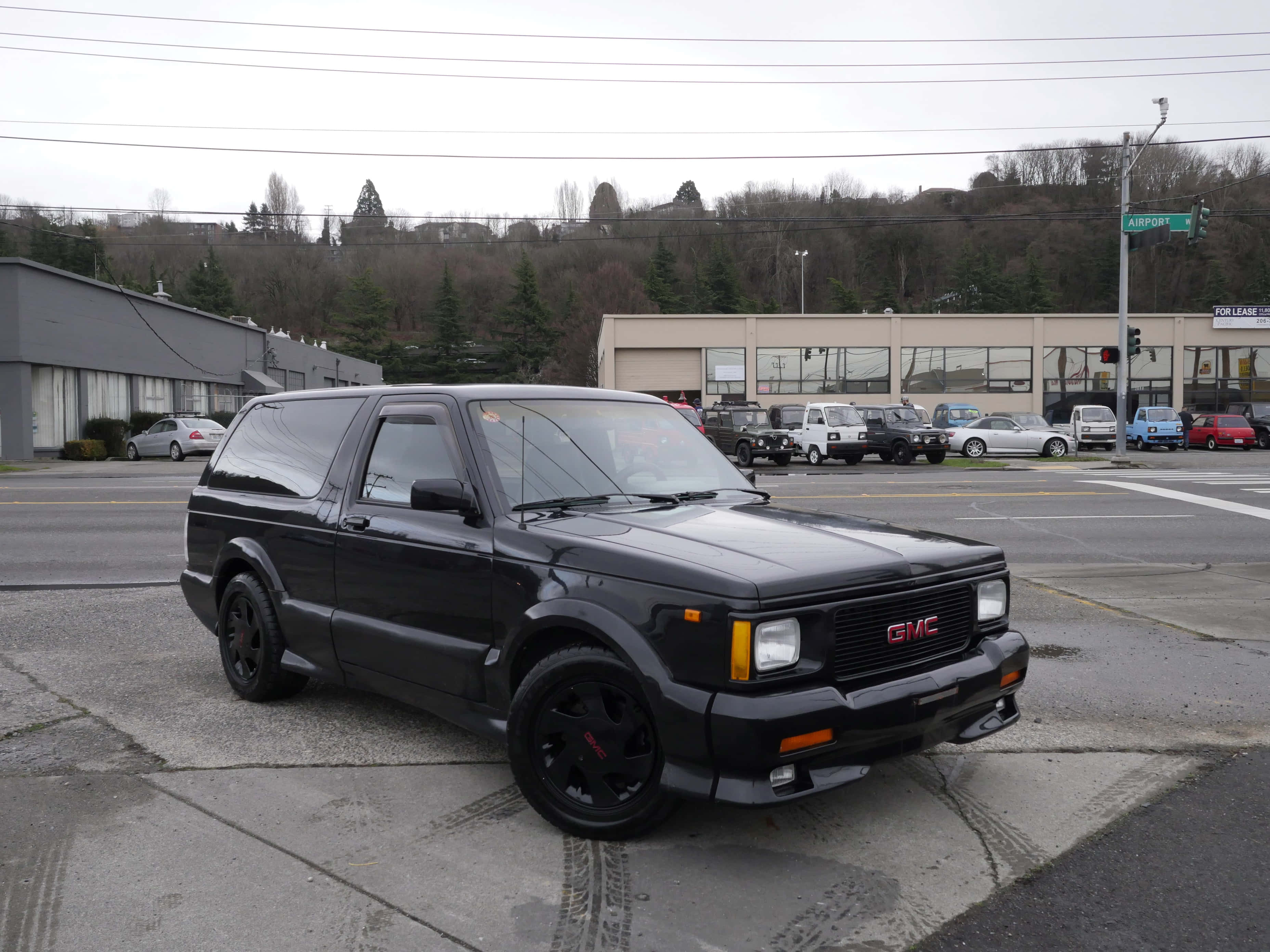 Gmc Typhoon 桌布 壁紙 — 一輛時尚的黑色GMC Safari SUV停泊在濕漉漉的城市街道上，背景有其他汽車和建築物。