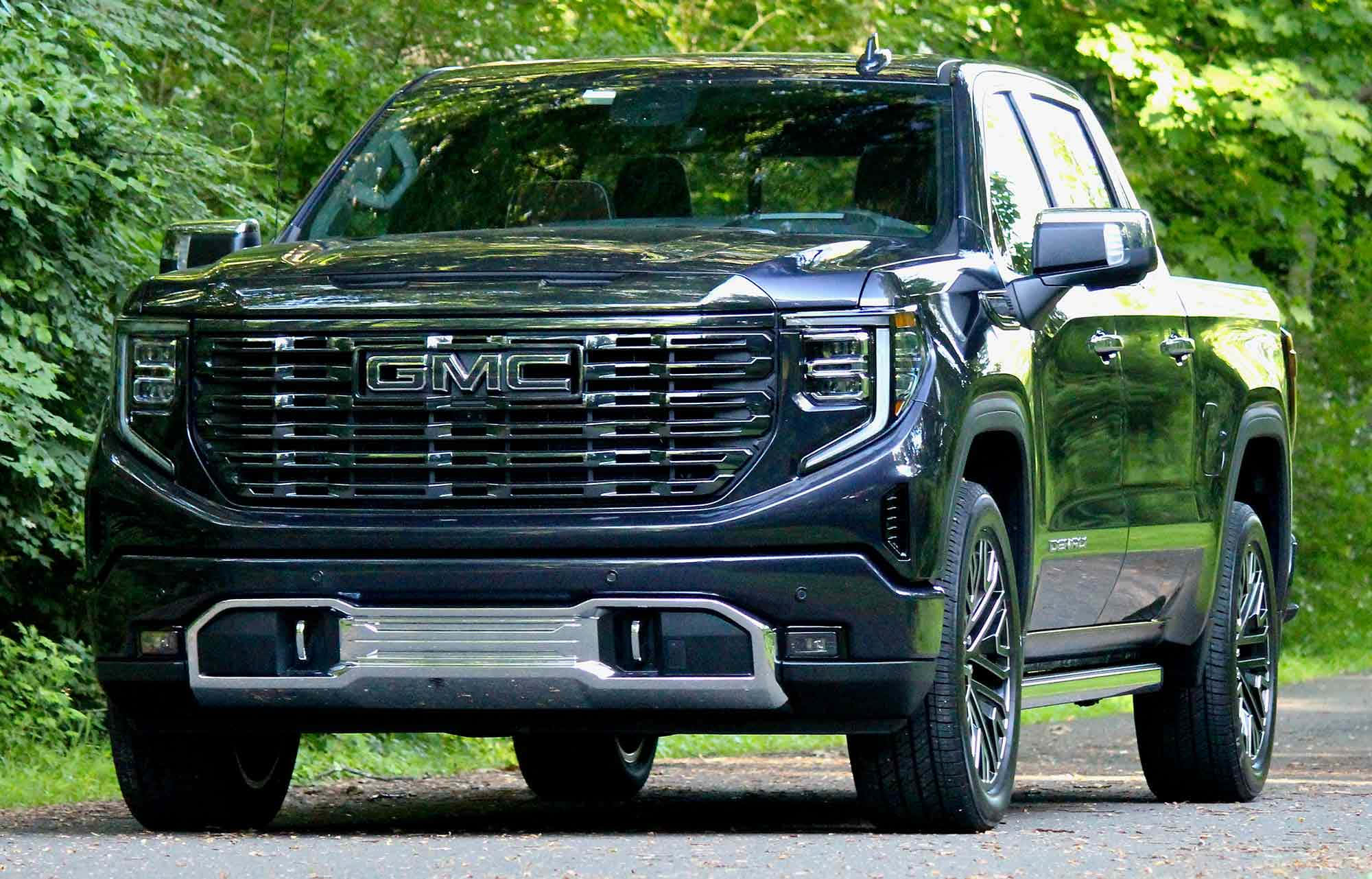 Gmc Sierra 桌布 壁紙 — 一架時尚的黑色GMC Sierra Denali皮卡車停泊在碎石路上，周圍是茂密的綠色森林。