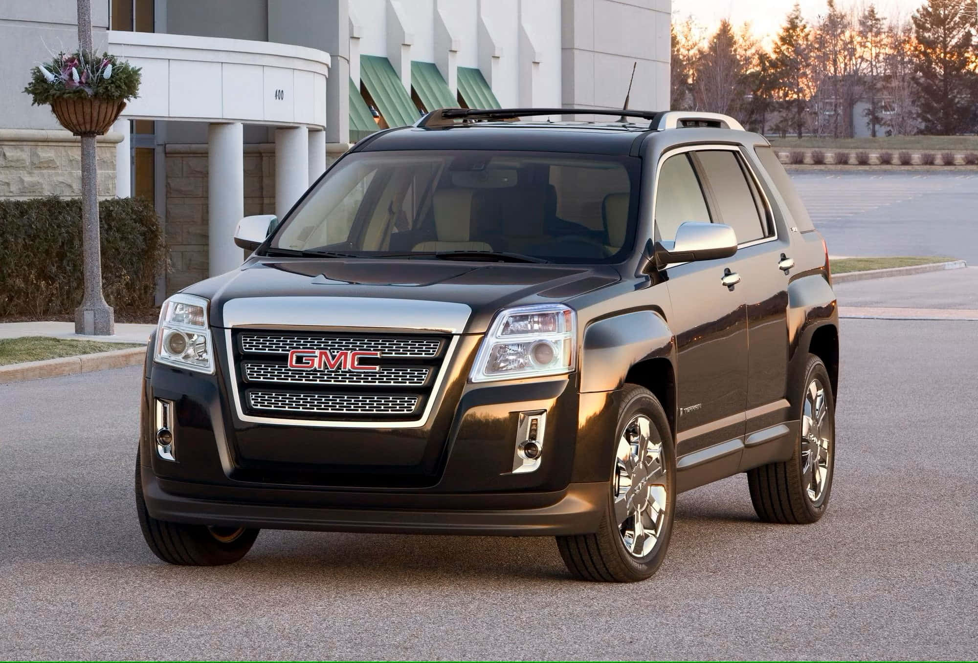 Gmc Terrain 桌布 壁紙 — 一架時尚的黑色GMC Terrain SUV停泊在鋪砌的停車場上，背景是現代建築和樹木，正值黃金時刻。