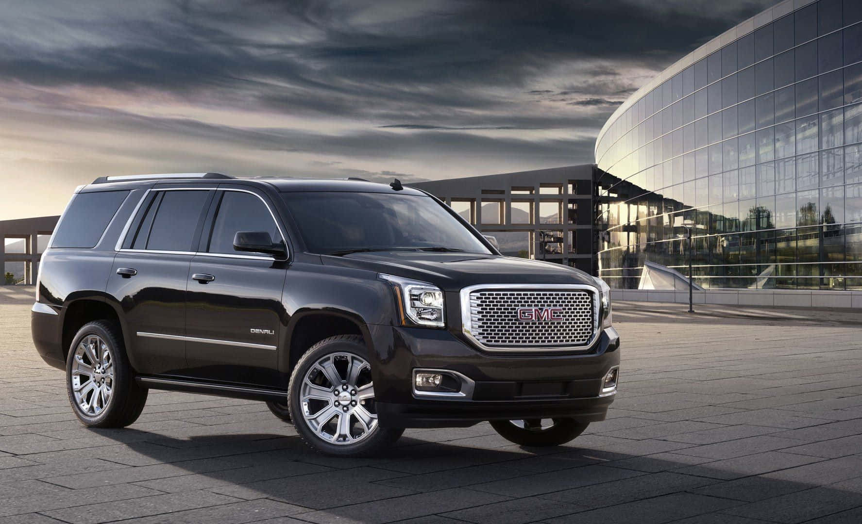 Gmc Yukon 桌布 壁紙 — 一輛時尚的黑色GMC Yukon SUV停泊在現代玻璃建築前的瓷磚廣場上，天空佈滿戲劇性的雲層。