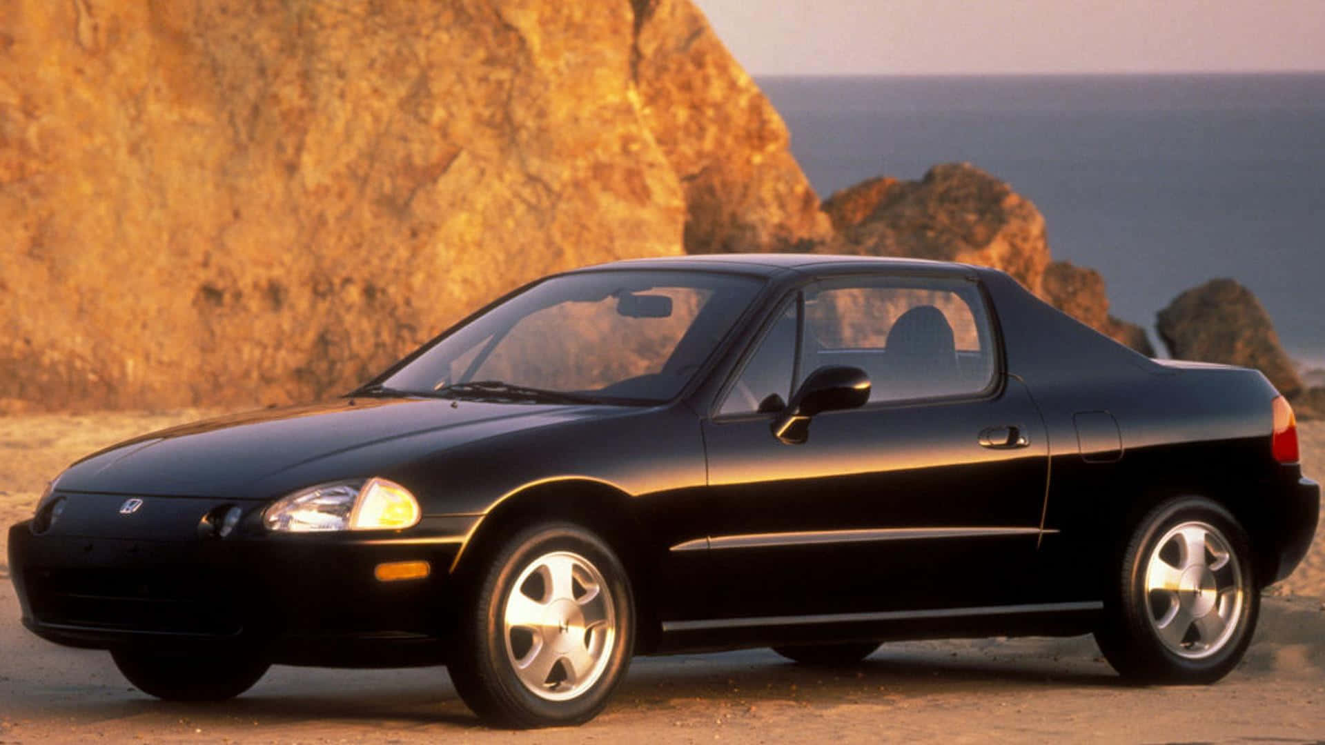 Honda Civic Del Sol 桌布 壁紙 — 一輛時尚的黑色本田S2000跑車停在日落時分的海岸懸崖上，溫暖的金色光線反射在其光滑的表面。