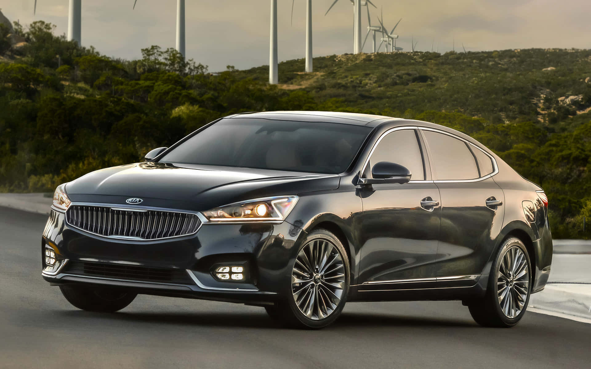 Kia Cadenza 桌布 壁紙 — 一輛時尚的黑色Kia轎車在柔和天空下，沿著一條有風力發電機和綠色山丘的道路行駛。