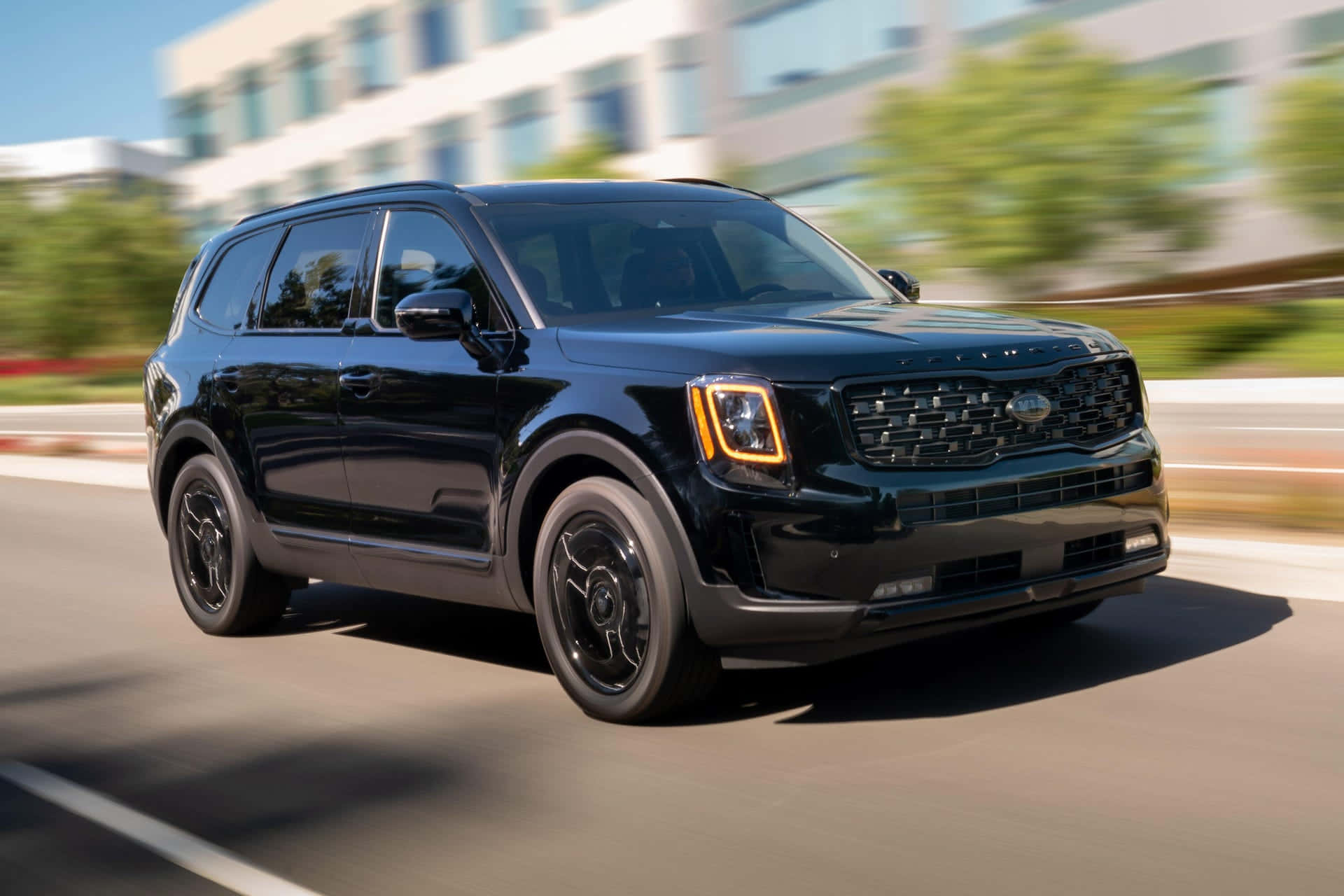 Kia Telluride 桌布 壁紙 — 一輛時尚的黑色Kia SUV在陽光明媚的城市道路上飛馳，樹木和建築物呈現動態模糊效果。