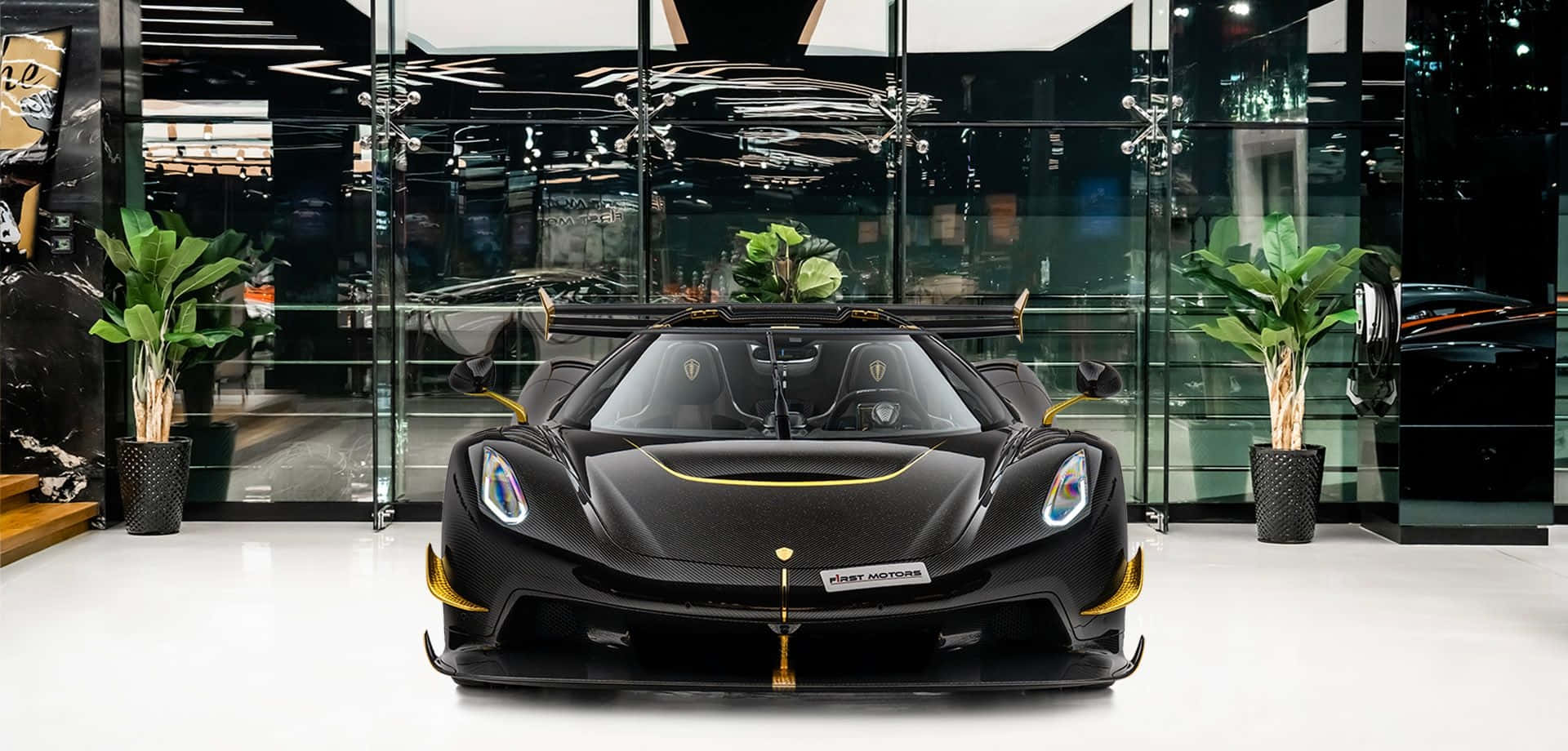  壁紙 — 一輛帶金色裝飾的時尚黑色Koenigsegg超跑，在現代玻璃陳列室中展示，兩旁擺放著盆栽植物。
