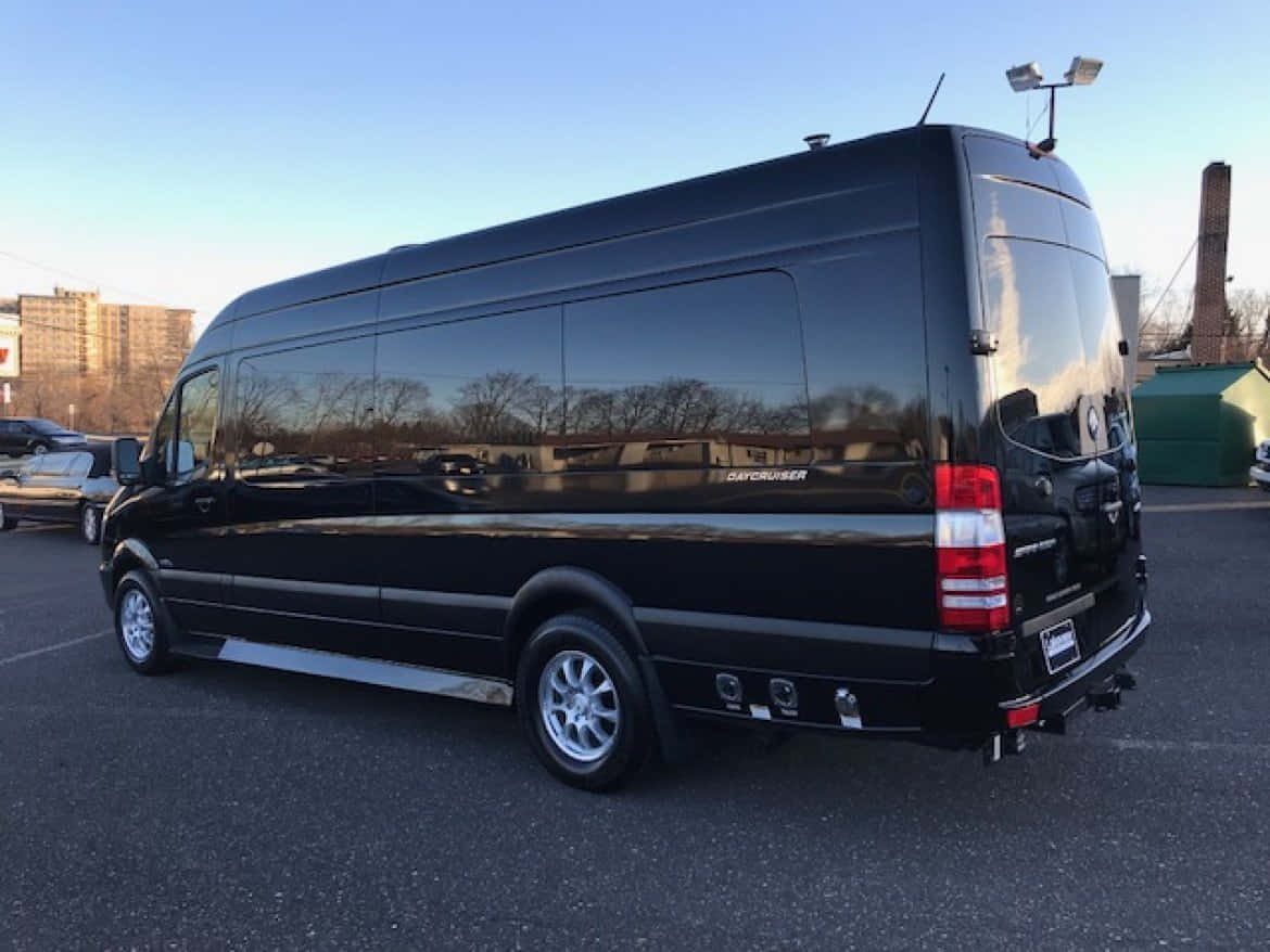 Mercedes Benz Sprinter 桌布 壁紙 — 一架時尚的黑色豪華客貨車停泊在瀝青停車場上，天空清澈湛藍，光亮的車窗倒映著光禿的樹木。
