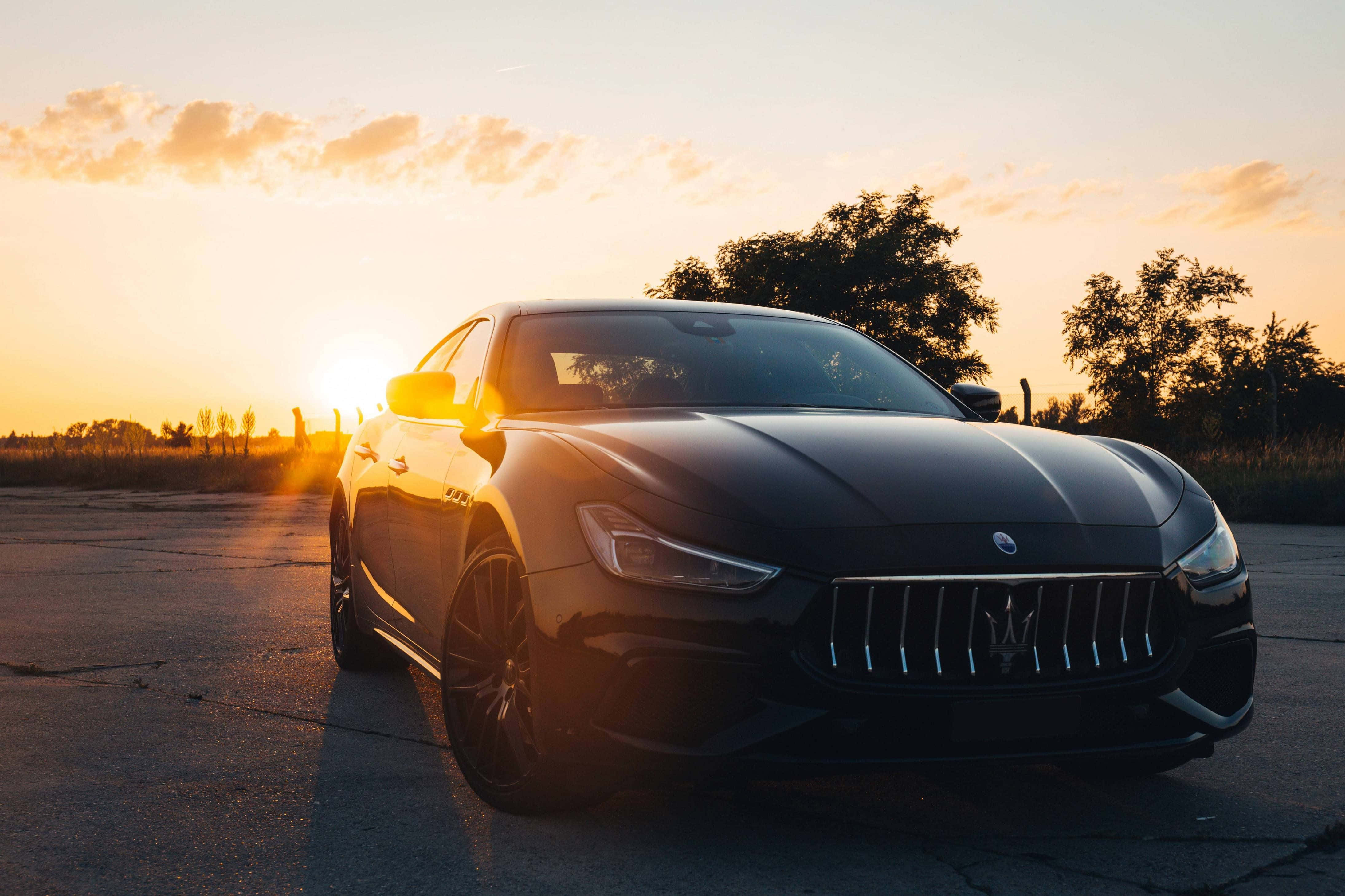 Maserati Ghibli 桌布 壁紙 — 一輛時尚的黑色瑪莎拉蒂跑車在金色夕陽下閃耀，背景是鋪砌的道路和樹木。