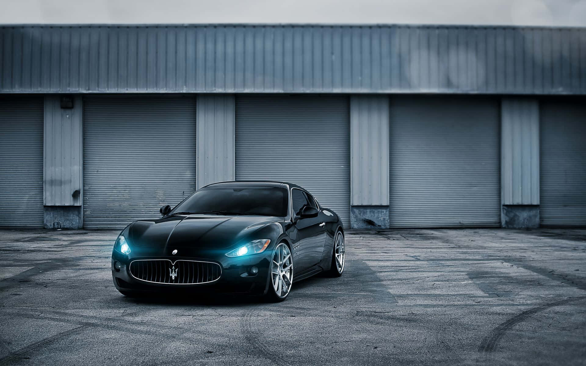 Luxury 桌布 壁紙 — 一輛時尚的黑色瑪莎拉蒂跑車，帶有發光的車頭燈，停在灰色工業建築前。