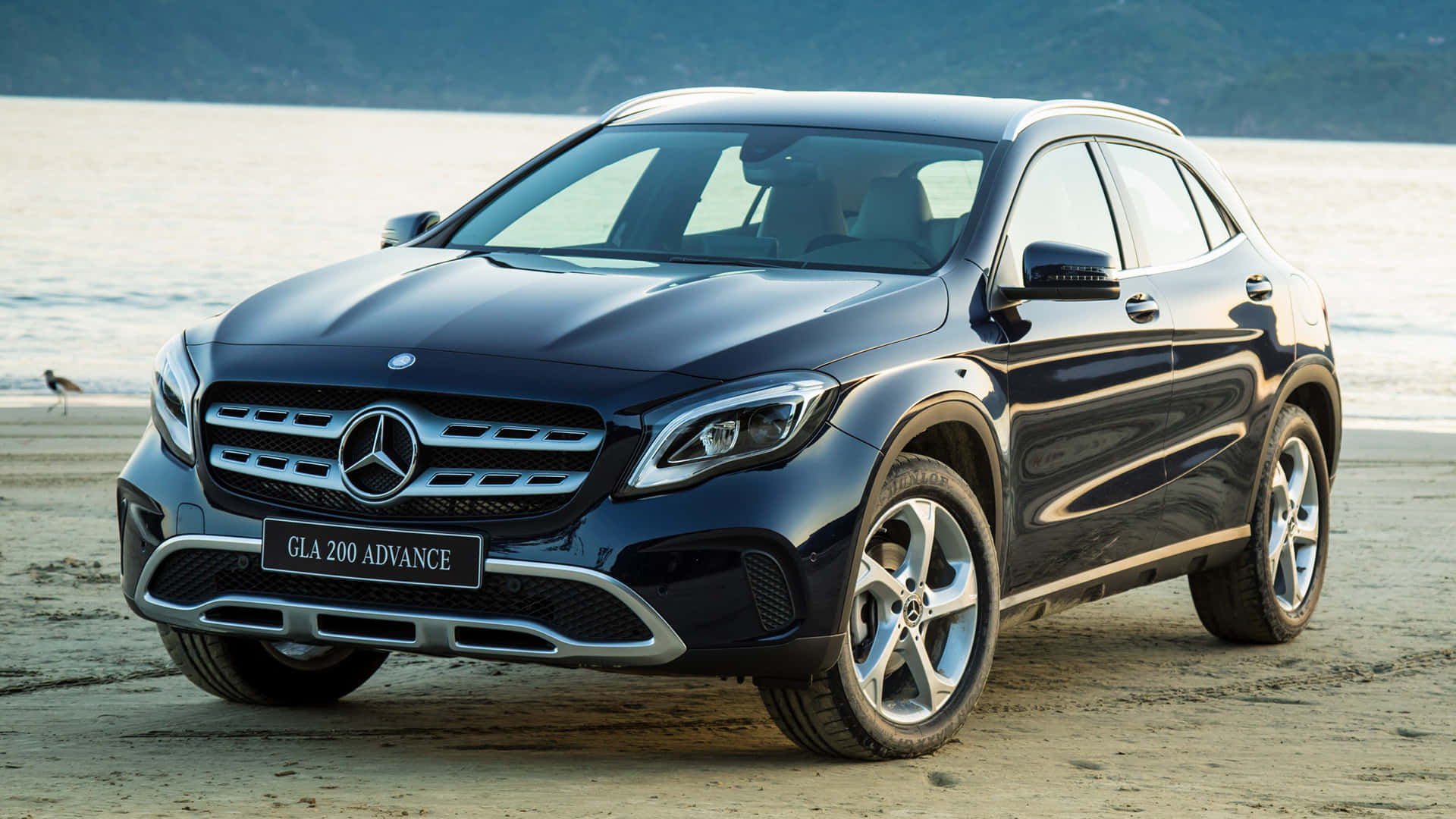 Mercedes Benz Gla-class 桌布 壁紙 — 一輛時尚的黑色 Mercedes-Benz GLA 200 Advance SUV 停泊在沙灘上，背景是海洋和山丘。