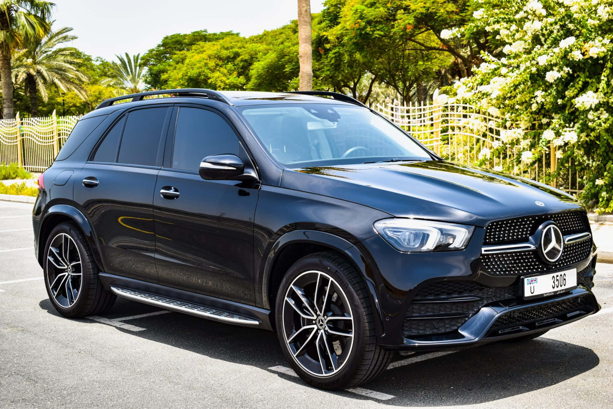 Mercedes Benz Gle-class 桌布 壁紙 — 一架時尚的黑色平治 GLE SUV 停泊在陽光普照的停車場，周圍環繞著茂密的綠樹和白色開花灌木。
