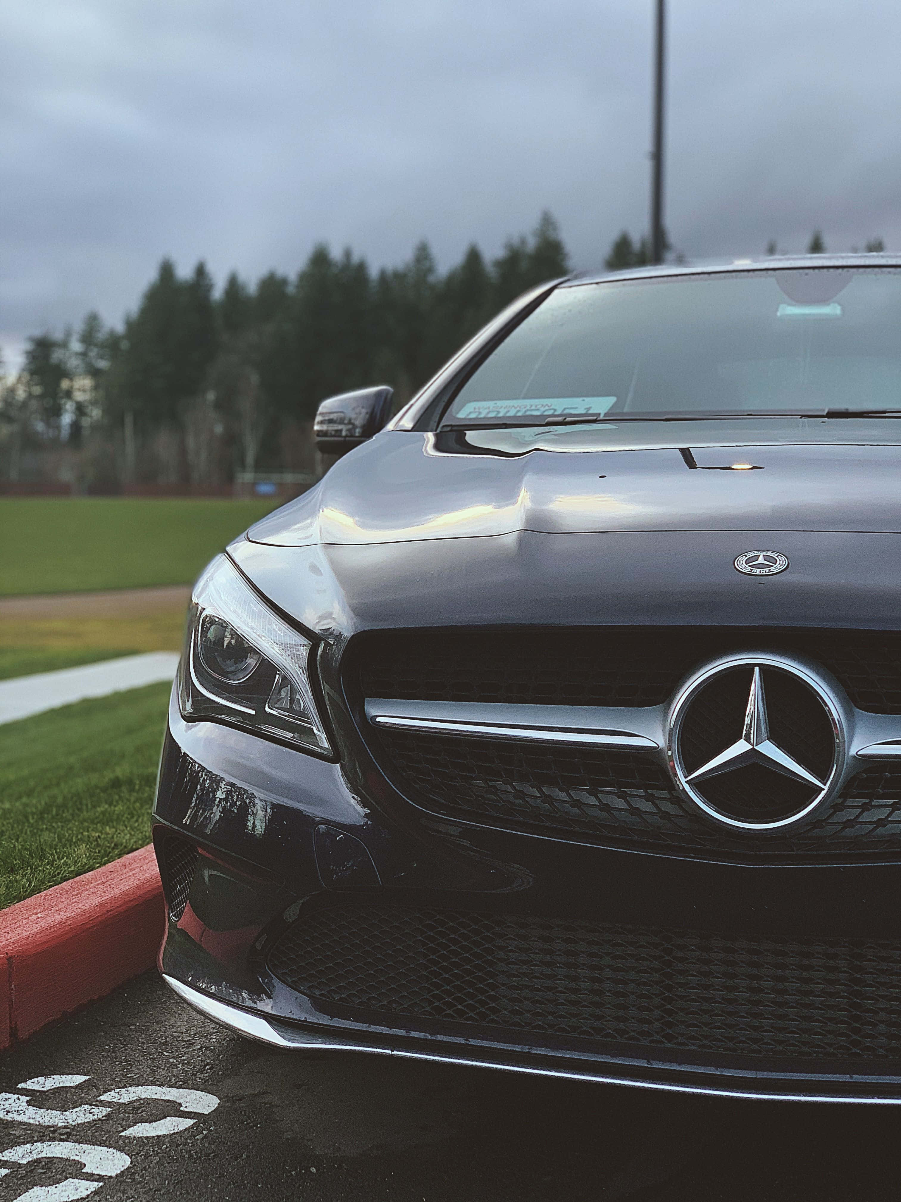 Mercedes Benz Cla-class 桌布 壁紙 — 一架時尚的黑色平治停泊在瀝青路上，旁邊是紅色路緣，背景是樹林，天空多雲。