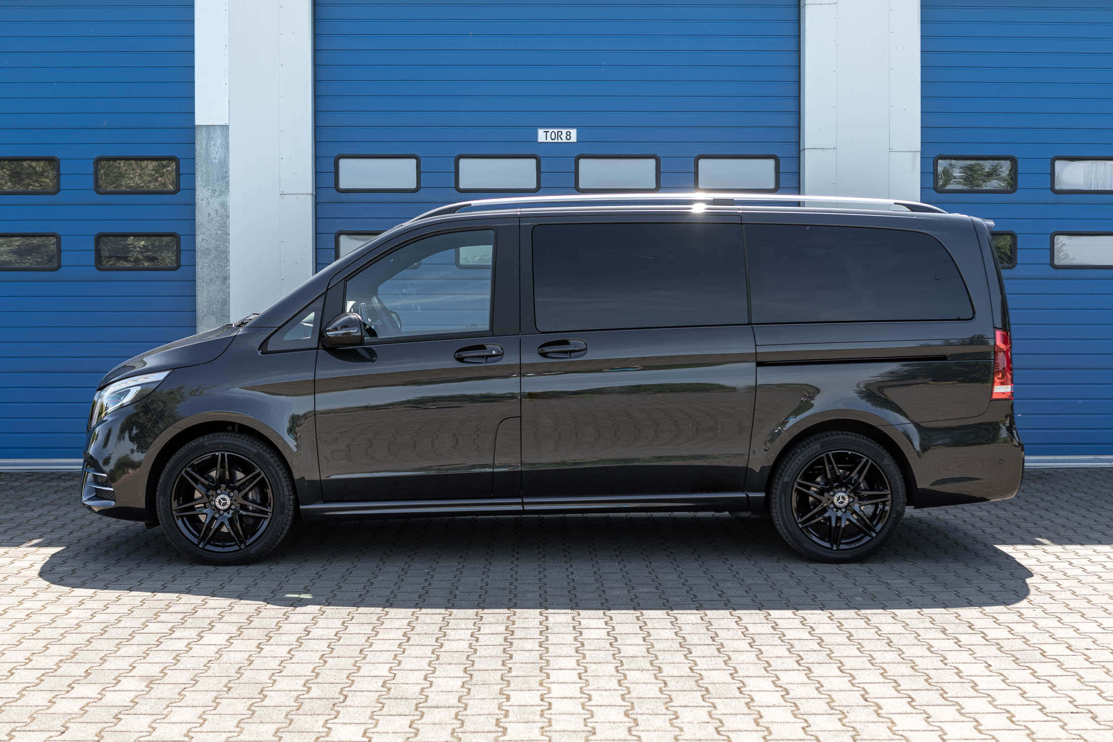 Mercedes Benz V-class 桌布 壁紙 — 一輛時尚的黑色 Mercedes-Benz V-Class 客貨車停泊在圖案行人路上，前面是充滿活力的藍色工業車庫門。