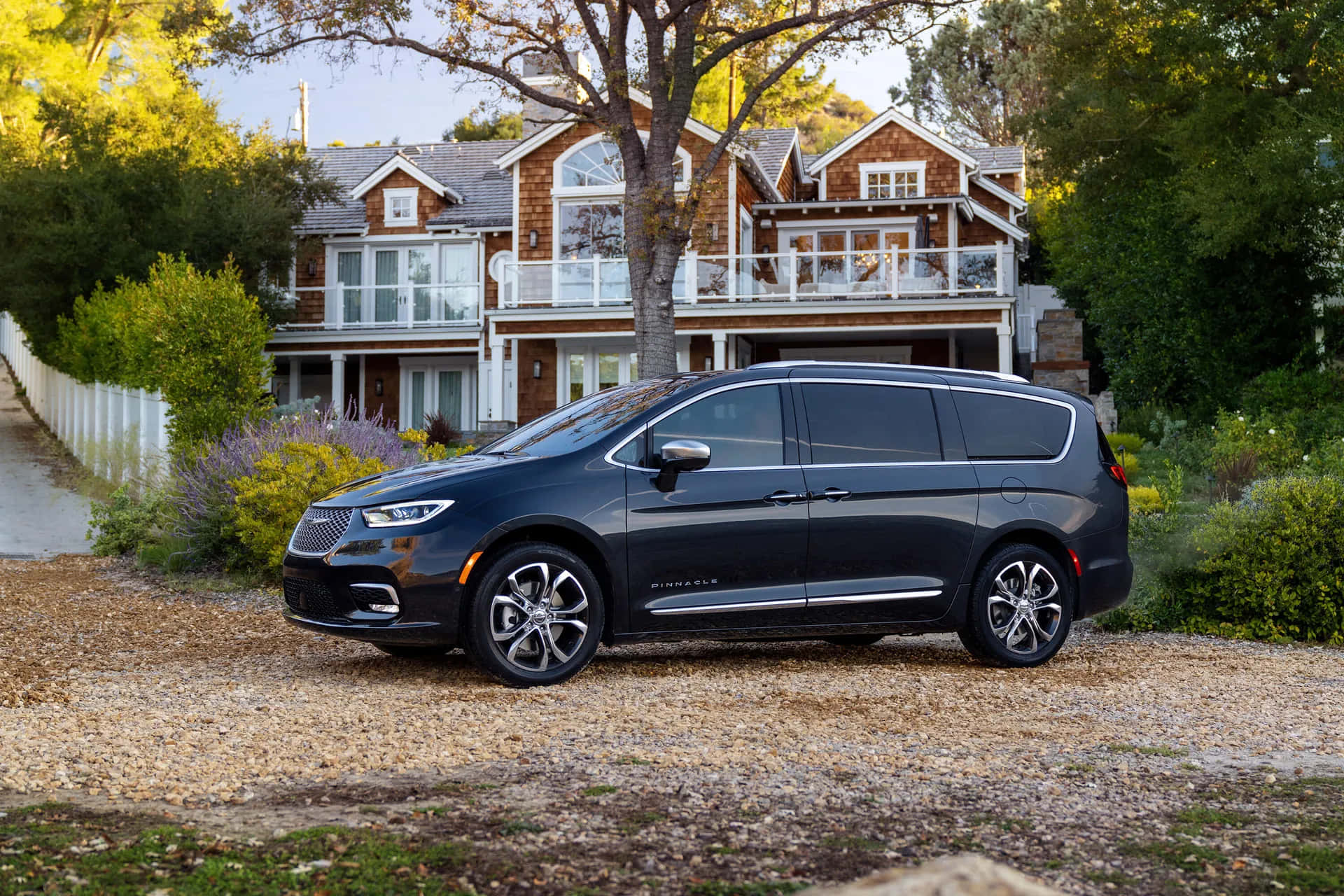 Chrysler Pacifica 桌布 壁紙 — 一輛時尚的黑色小型貨車停泊在碎石車道上，前面是一間被茂密綠樹環繞的大型木屋。