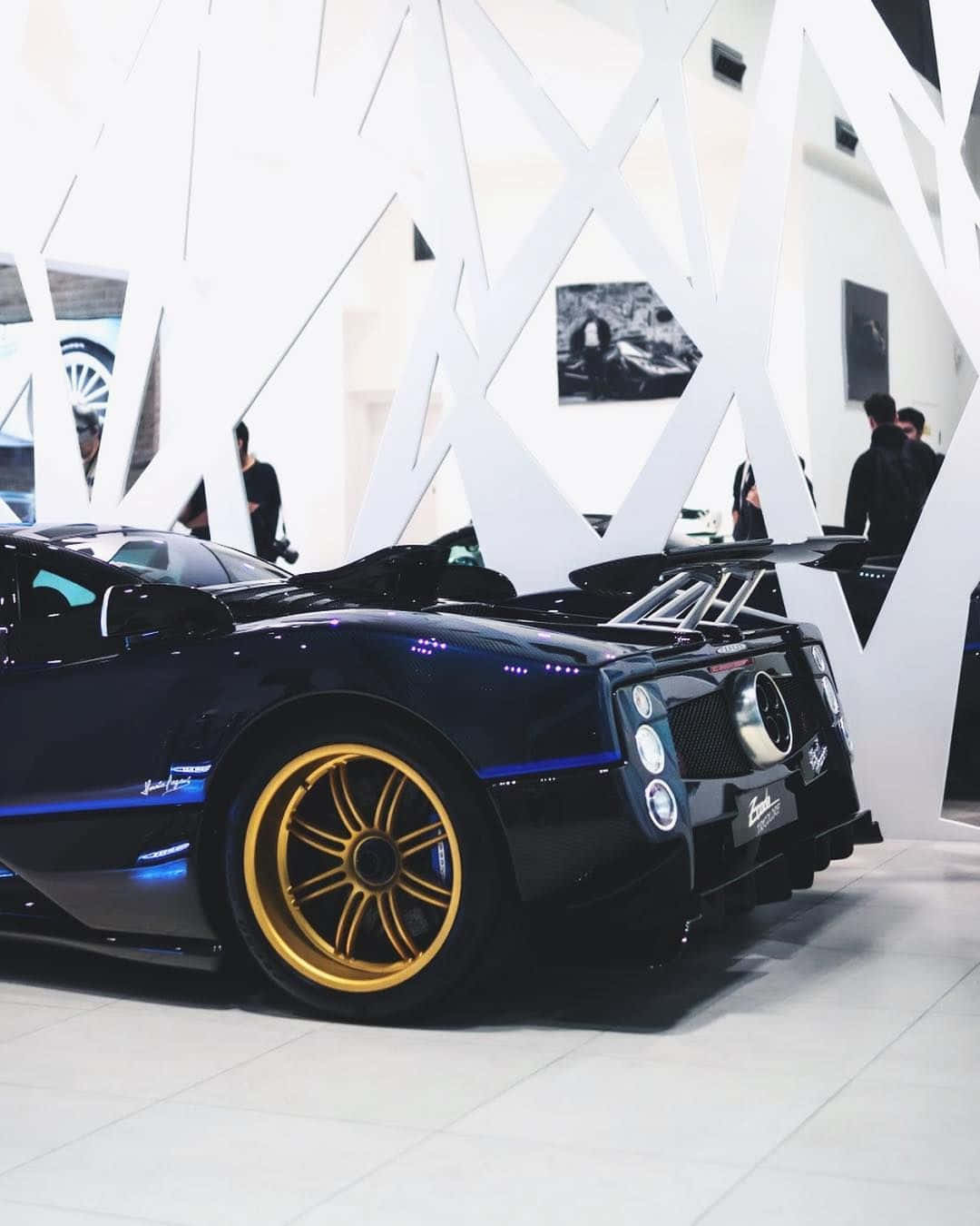 Pagani Zonda Tricolore 桌布 壁紙 — 一輛時尚的黑色帕加尼Huayra，配金色輪圈和藍色點綴，在現代白色畫廊的明亮燈光下閃閃發光。
