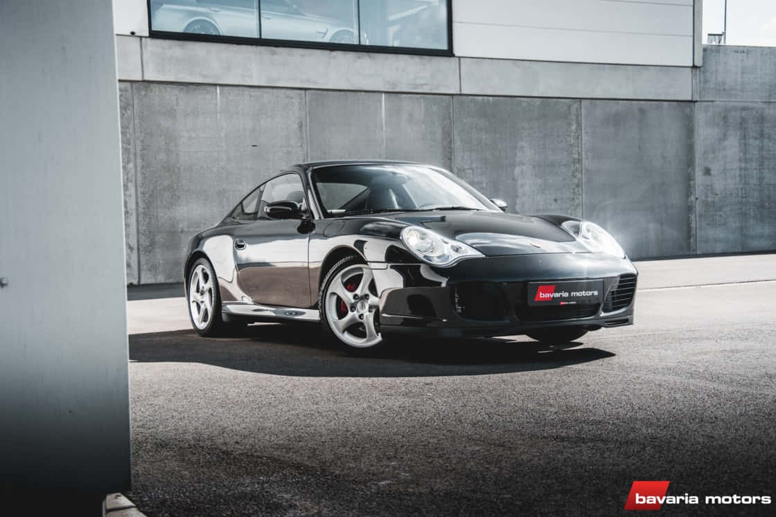 Porsche 996 桌布 壁紙 — 一輛時尚的黑色保時捷911跑車在明亮的日光下停靠在現代混凝土建築旁閃閃發光。