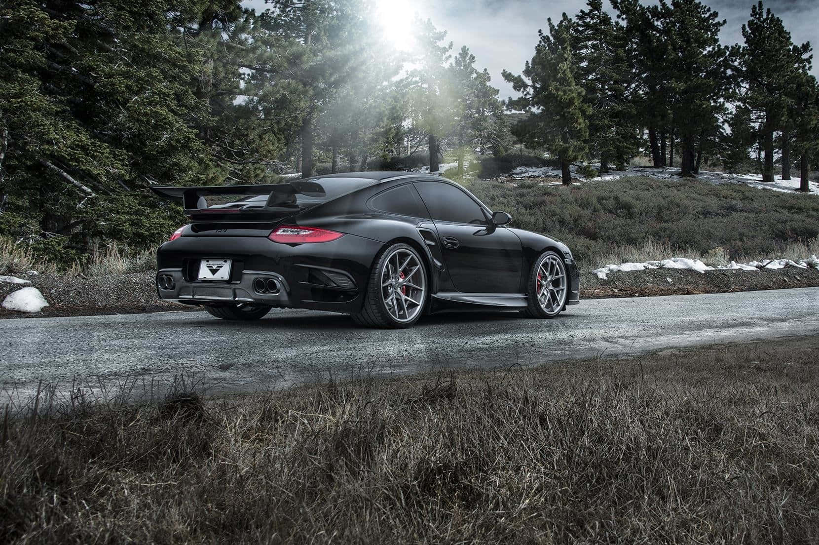 Porsche 997 桌布 壁紙 — 一輛時尚的黑色保時捷跑車，帶有大型後擾流板，停泊在森林道路上，陽光穿過松樹照射下來。