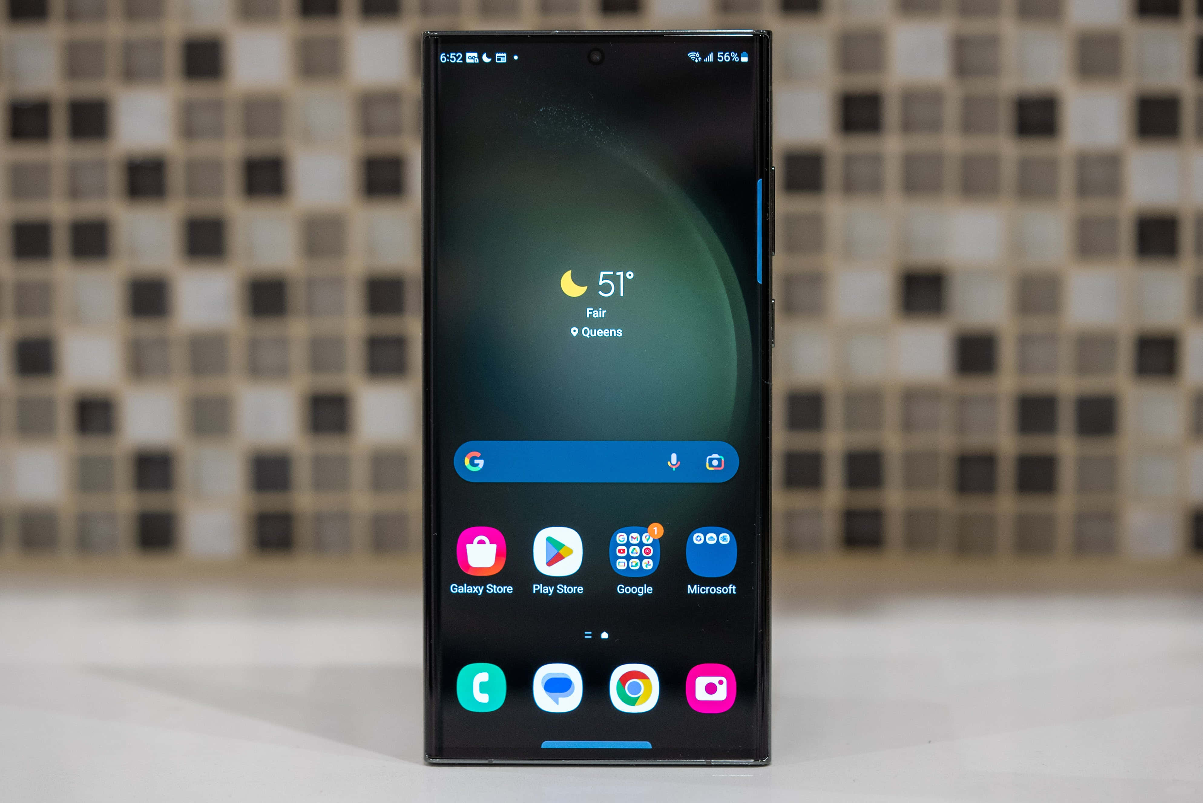 Samsung S23 Ultra 桌布 壁紙 — 一部時尚的黑色智能手機在模糊的馬賽克瓷磚背景上顯示天氣小工具和應用程式圖標。
