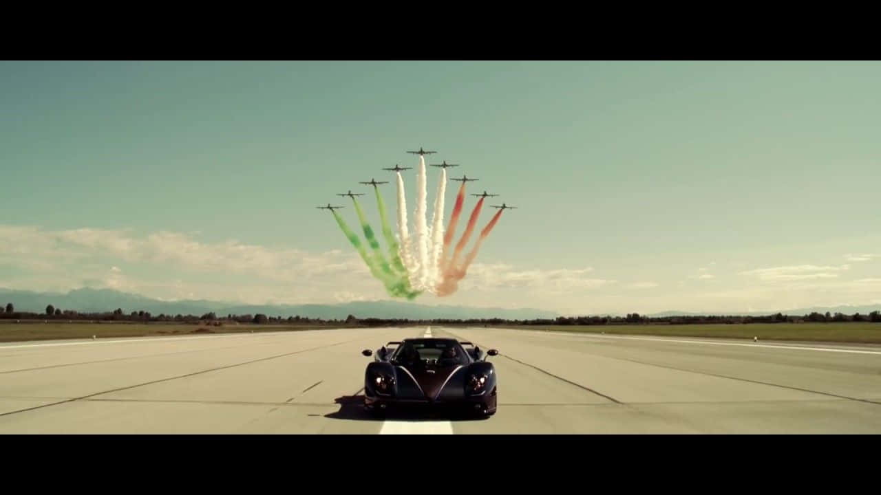 Pagani Zonda Tricolore 桌布 壁紙 — 一架流線型黑色跑車面向一隊噴射機，噴射機在機場跑道上空釋放出綠色、白色和紅色的煙霧軌跡。