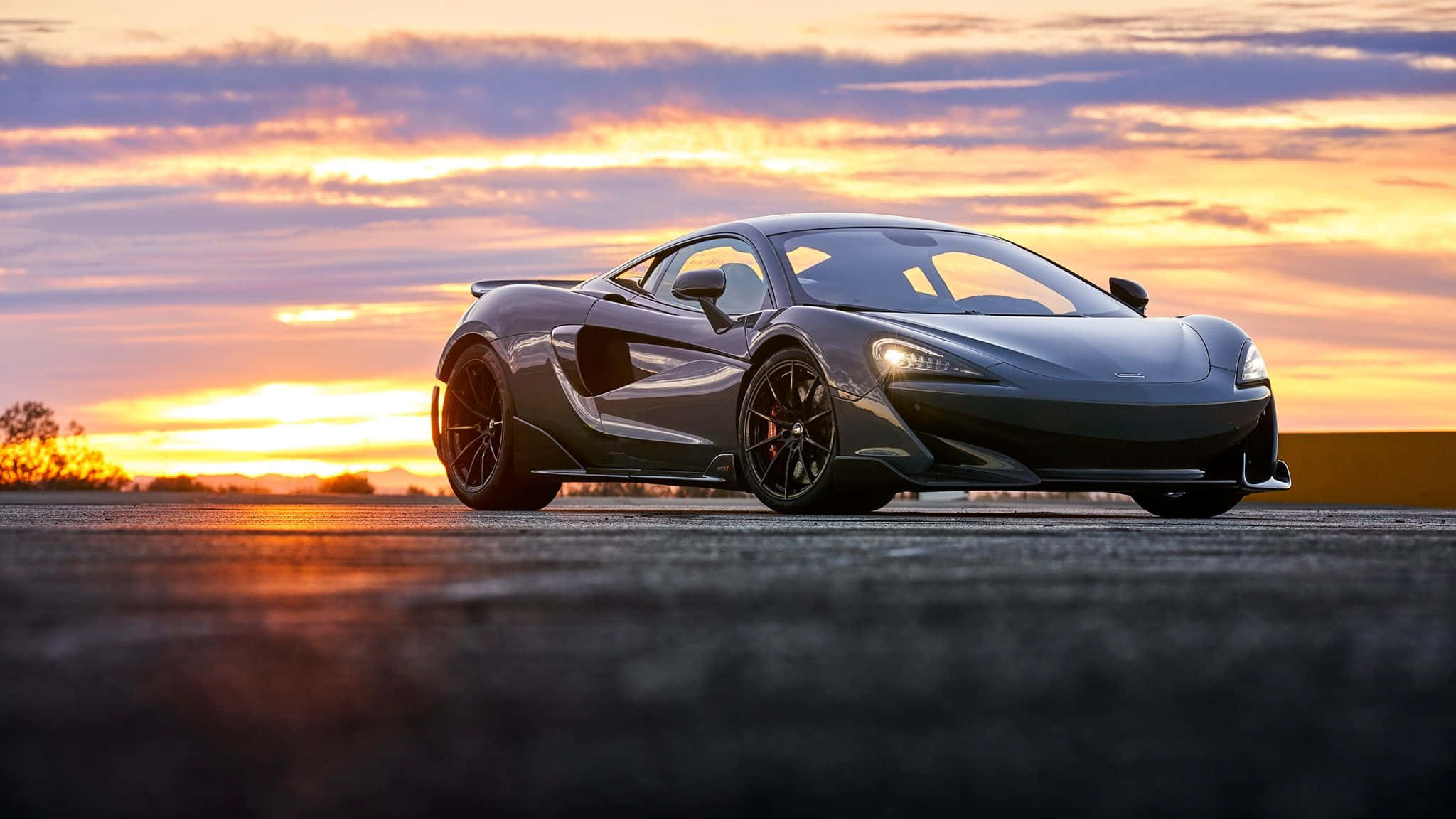 Mclaren 600lt 桌布 壁紙 — 一輛時尚的黑色跑車在充滿活力的日落天空下閃耀，天空中有橙色和紫色的雲彩。