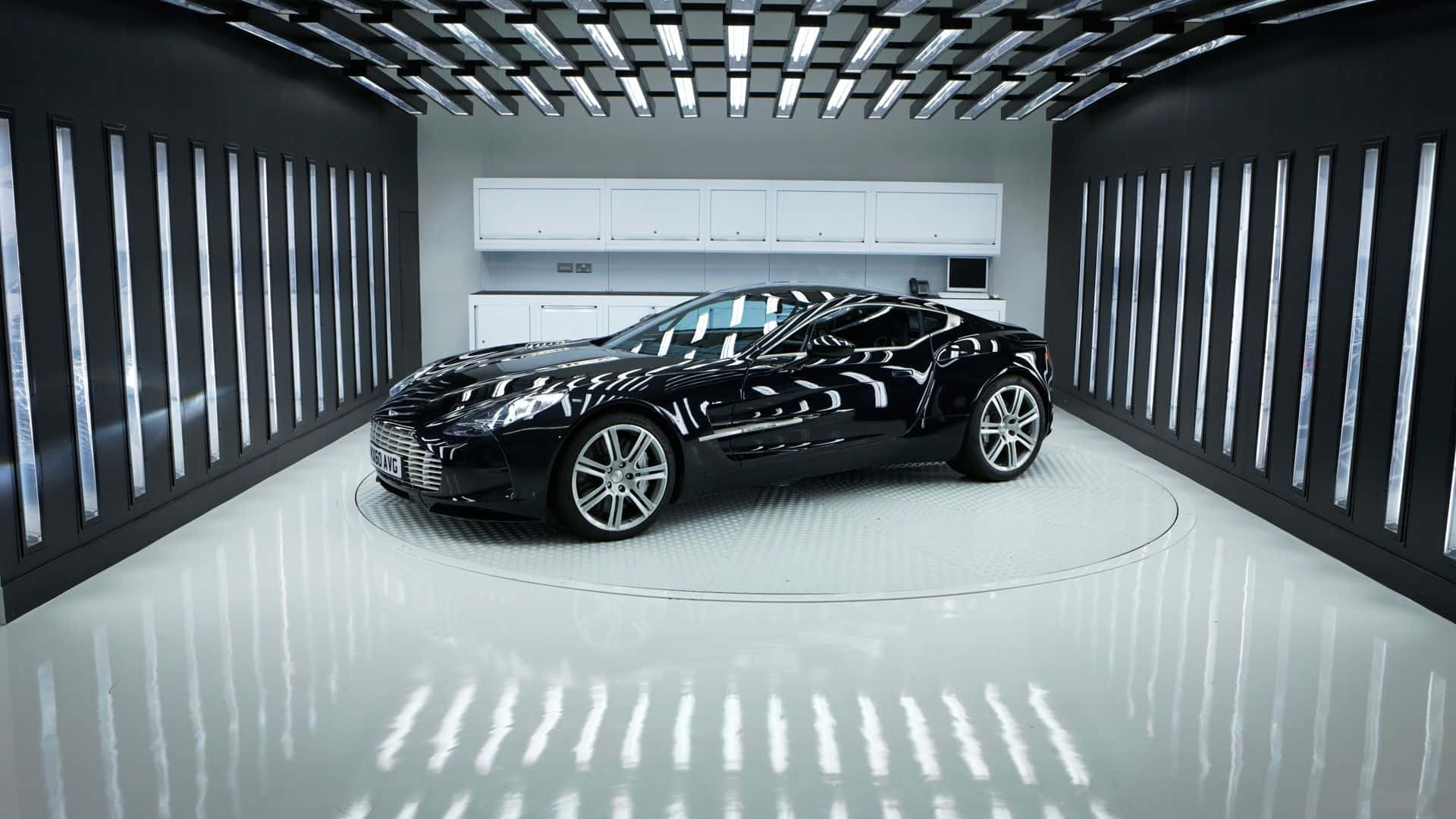 Aston Martin One-77 桌布 壁紙 — 一輛時尚的黑色跑車在明亮的攝影棚燈光下，於一個被垂直燈板包圍的圓形平台上閃閃發光。