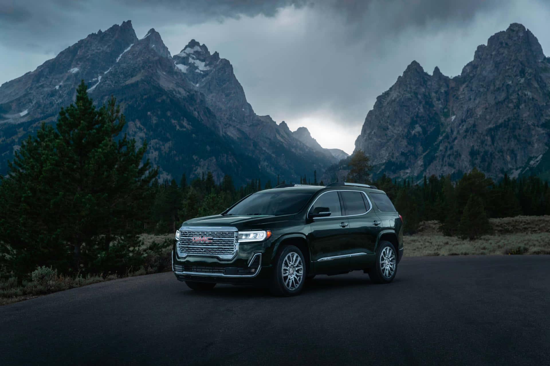Gmc Acadia 桌布 壁紙 — 一輛時尚的黑色SUV停泊在道路旁，背景是戲劇性的多雲天空下高聳的積雪山脈。