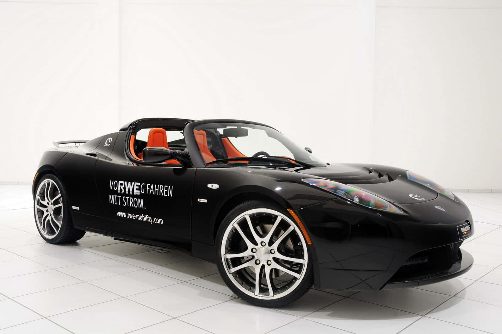 Tesla Roadster 桌布 壁紙 — 一架時尚的黑色Tesla Roadster開篷車，配橙色座椅和白色輪圈，停放在明亮的白色瓷磚工作室中。