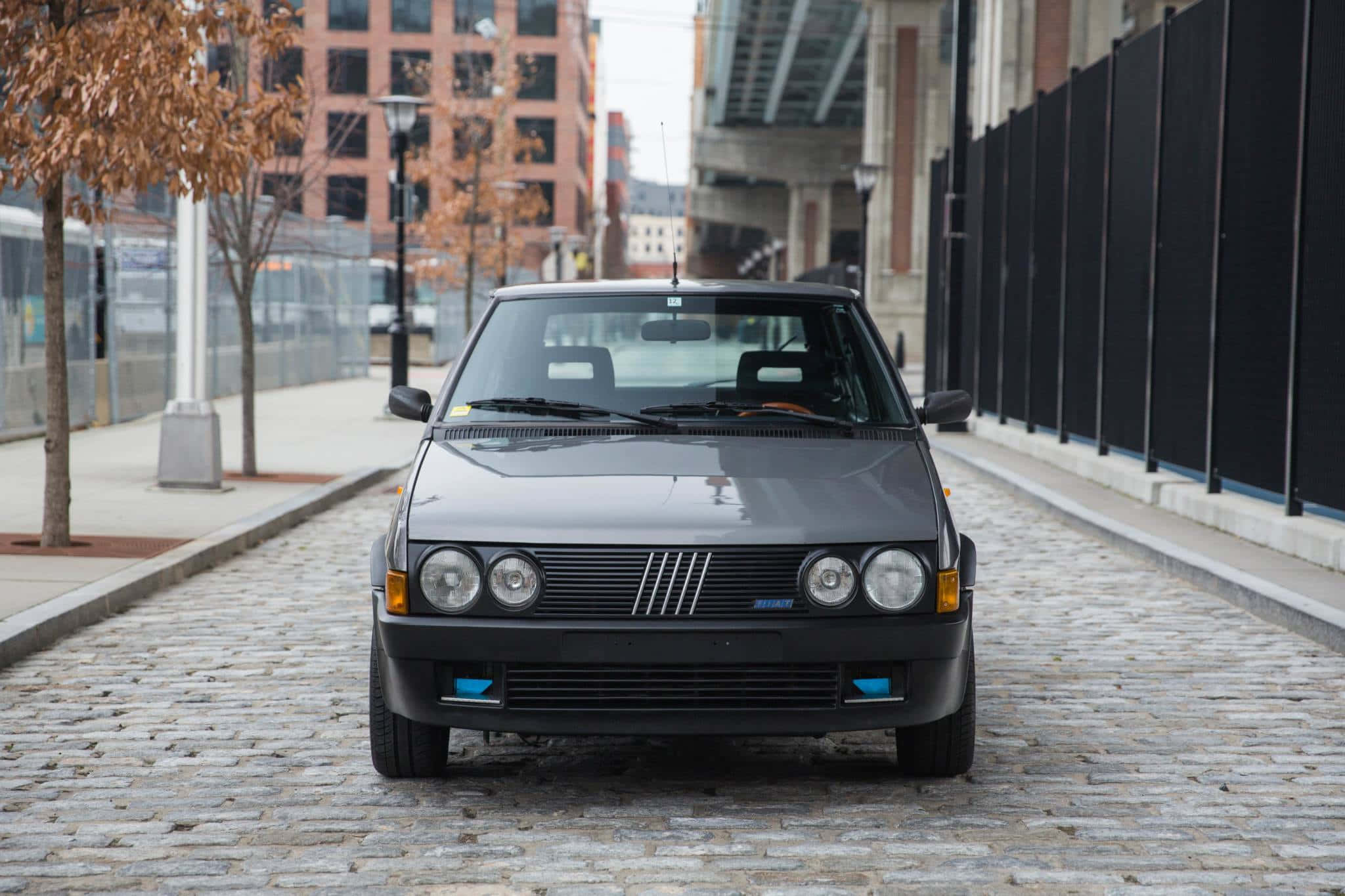 Fiat Ritmo 桌布 壁紙 — 一輛時尚的黑色復古車停在鵝卵石街道上，街道兩旁種滿了秋天的樹木和城市建築。