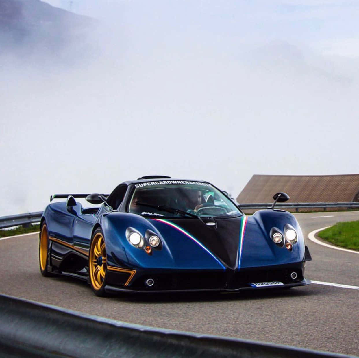 Pagani Zonda Tricolore 桌布 壁紙 — 一輛時尚的藍黑配色帕加尼超級跑車配金色輪圈，在迷霧繚繞的蜿蜒山路上行駛。