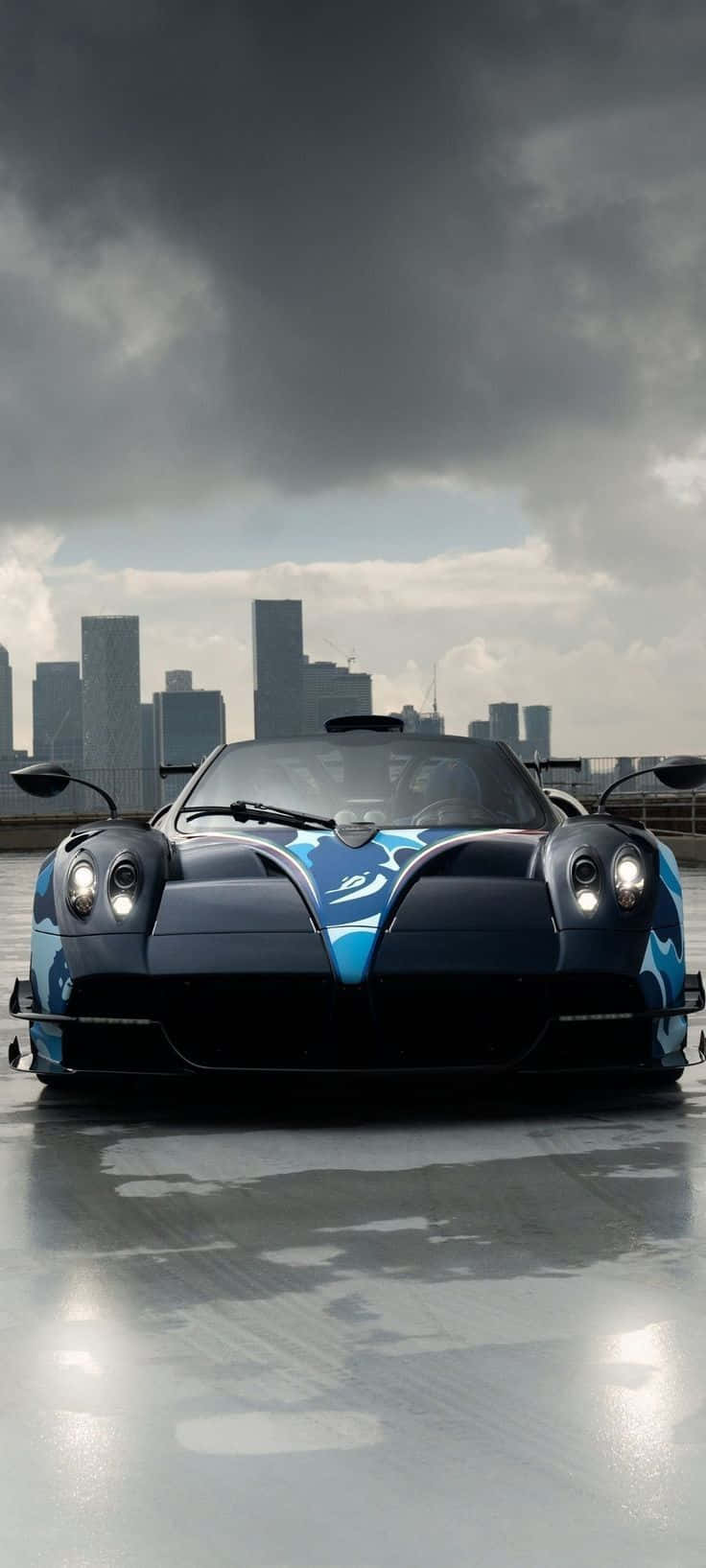 Pagani Huayra Roadster Bc 桌布 壁紙 — 一輛時尚的藍黑色跑車，帶有發光的車頭燈，停在潮濕的反光表面上，背景是暴風雨天空下的城市天際線。