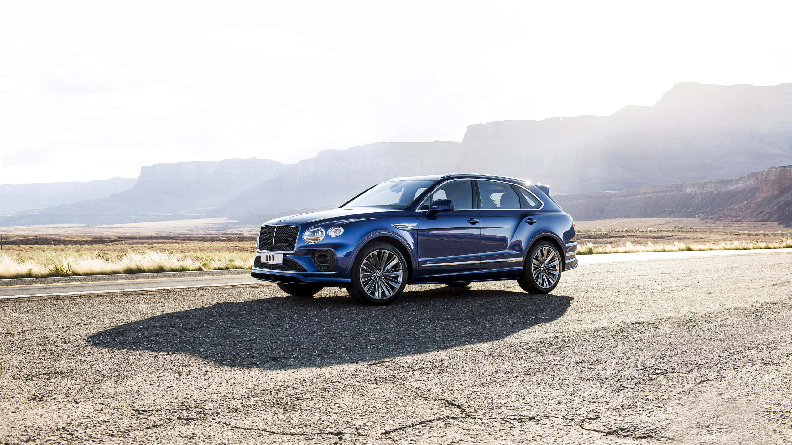Bentley Bentayga 桌布 壁紙 — 一輛時尚的藍色Bentley Bentayga SUV停泊在沙漠公路上，背景是崎嶇的山脈，在明亮的日光下。