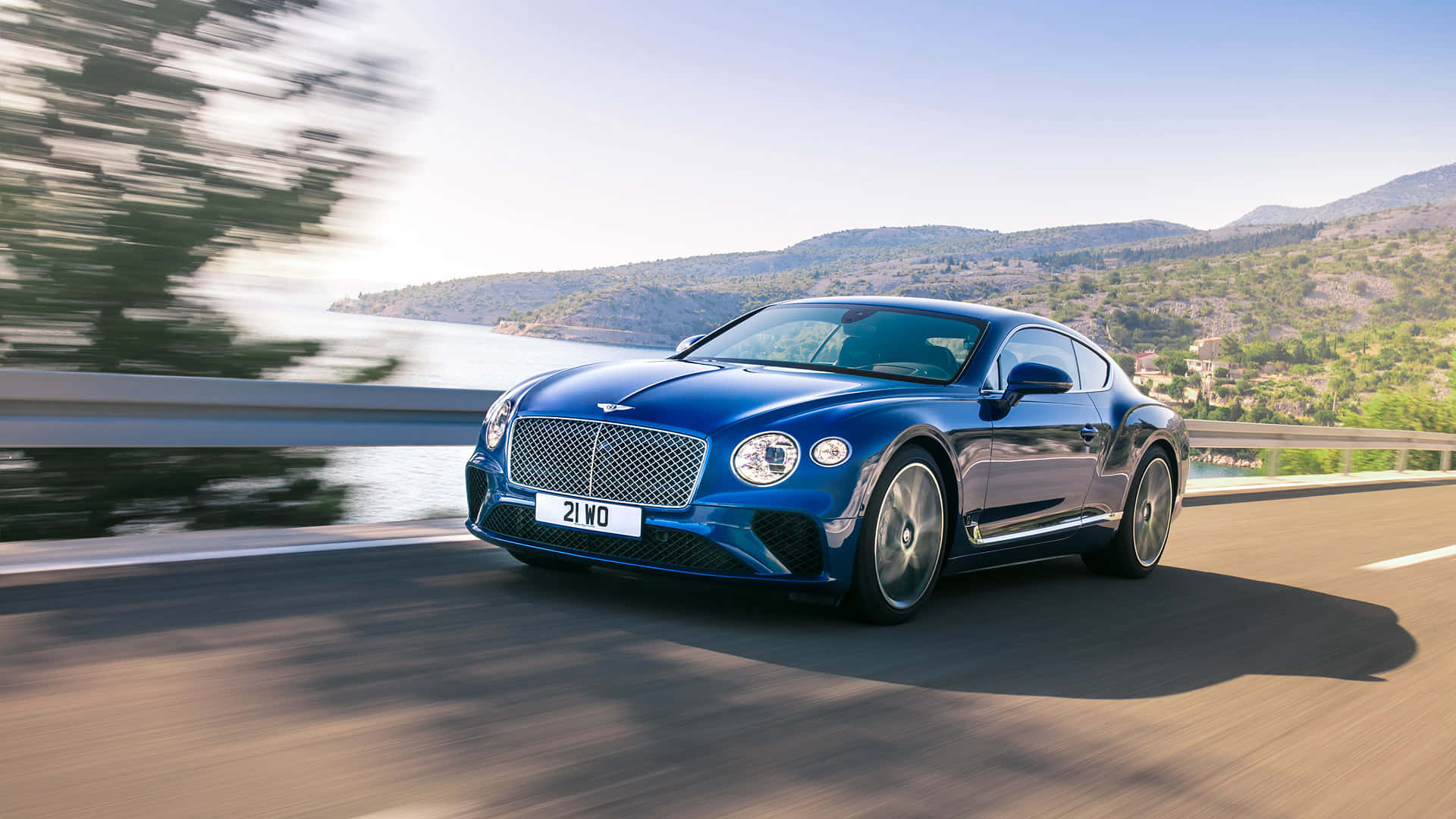 Bentley Continental Gt 桌布 壁紙 — 一輛時尚的藍色 Bentley Continental GT 沿著海岸公路飛馳，背景是模糊的綠色山丘和水面。
