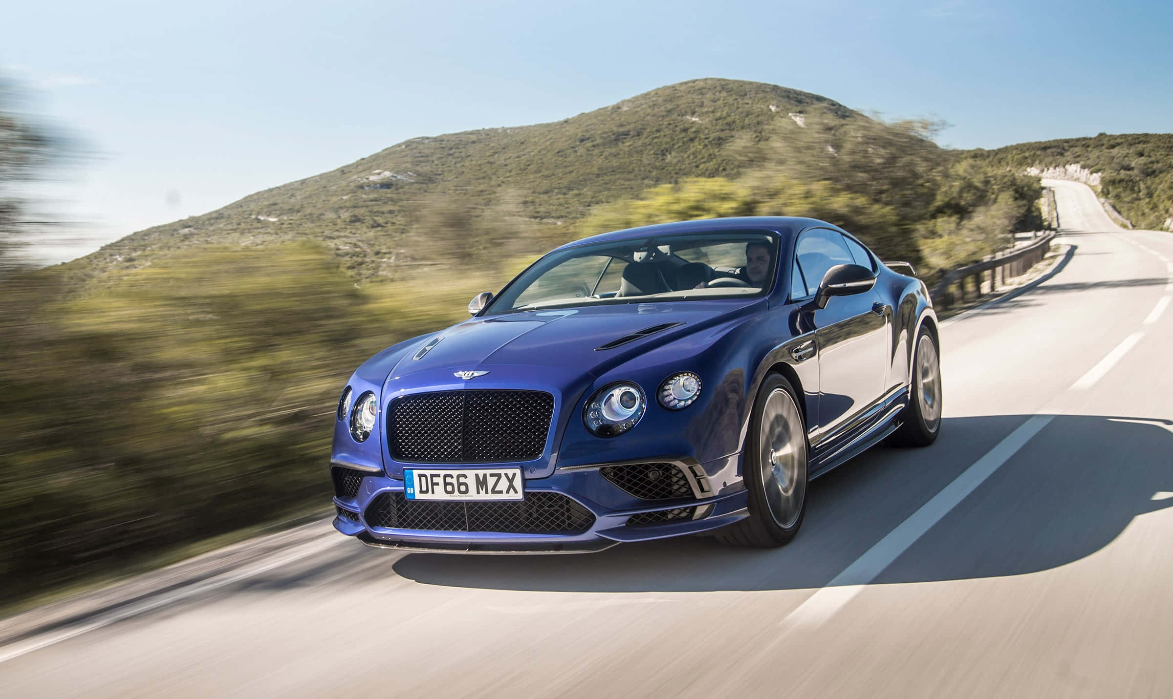 Bentley Continental Supersports 桌布 壁紙 — 一輛時尚的藍色 Bentley Continental GT 在晴朗的天空下，沿著蜿蜒的山路疾馳，周圍的綠樹和山丘呈現模糊效果。