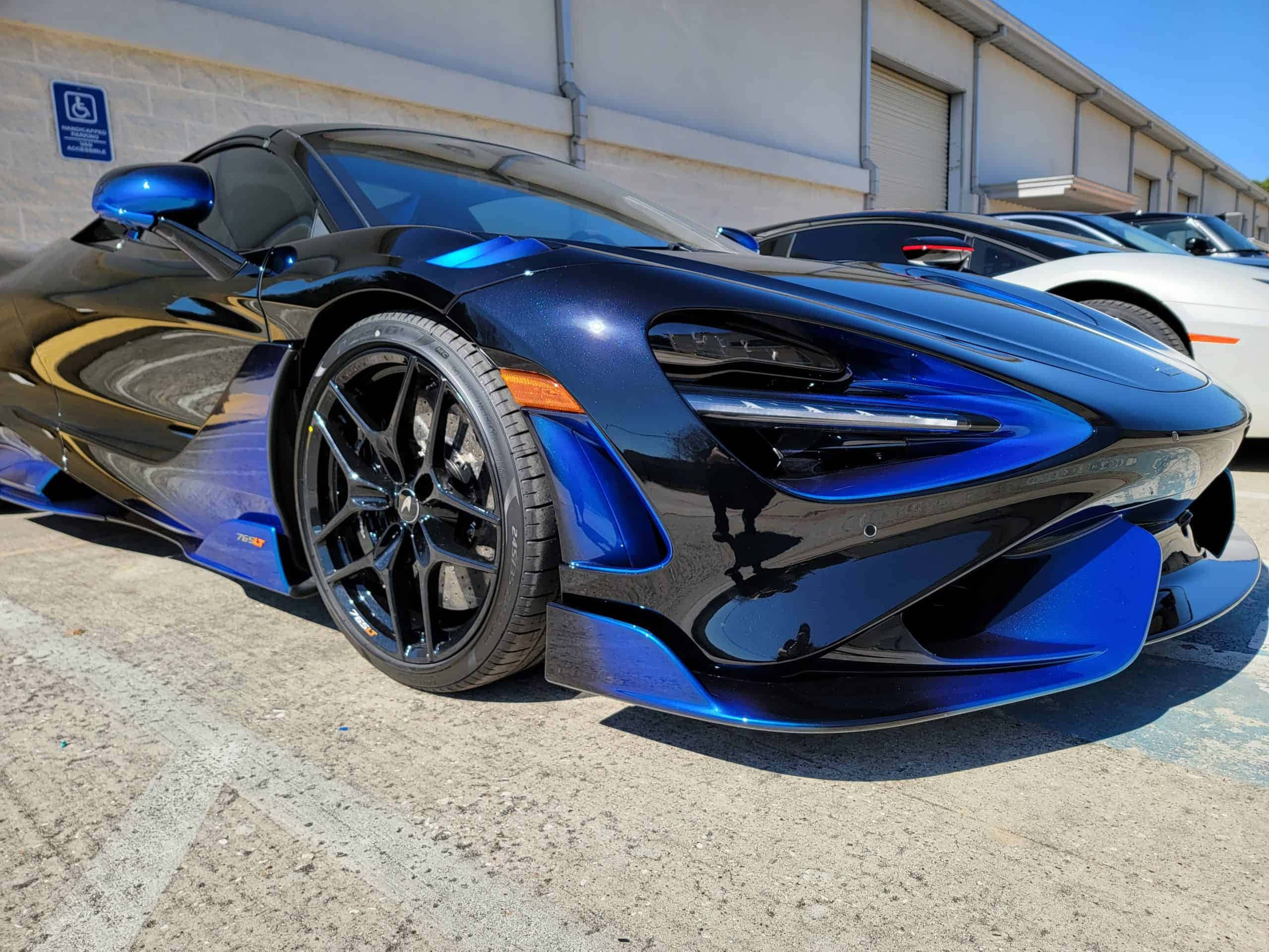 Mclaren 765lt 桌布 壁紙 — 一架時尚的藍黑McLaren跑車在明亮的陽光下閃閃發光，停泊在工業區。