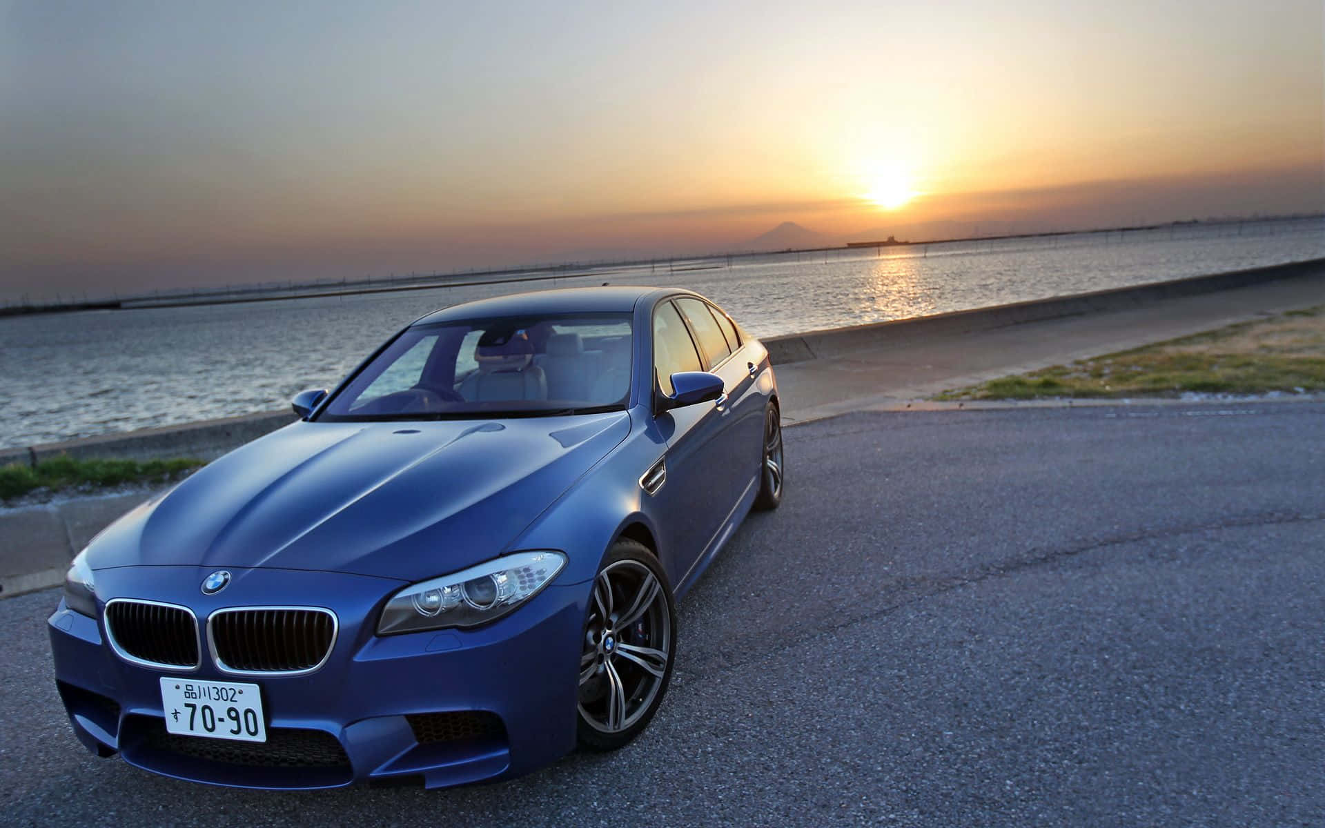 Bmw M5 桌布 壁紙 — 一架時尚的藍色BMW M5停在海濱道路上，夕陽在水面上投下金色光芒。