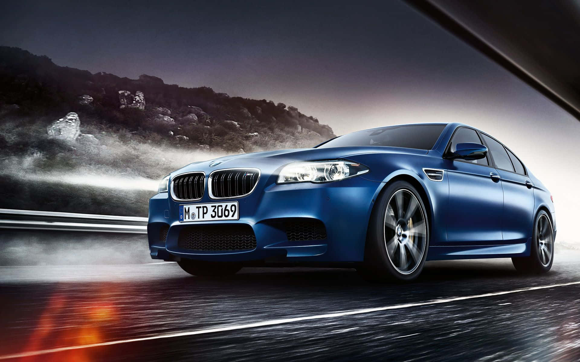 Bmw M5 桌布 壁紙 — 一輛時尚的藍色BMW M5在夜間濕滑的道路上飛馳，霧氣升騰，閃亮的車頭燈劃破黑暗。
