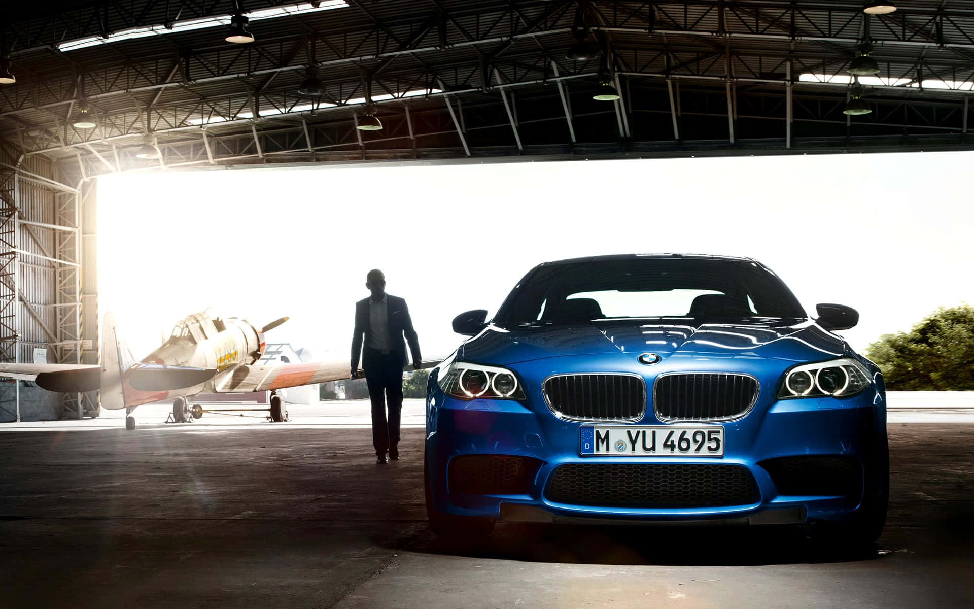 Bmw M5 桌布 壁紙 — 一架時尚的藍色BMW跑車在機庫內明亮燈光下閃閃發光，一名身穿西裝的男子正沿著一架復古飛機旁邊走向它。