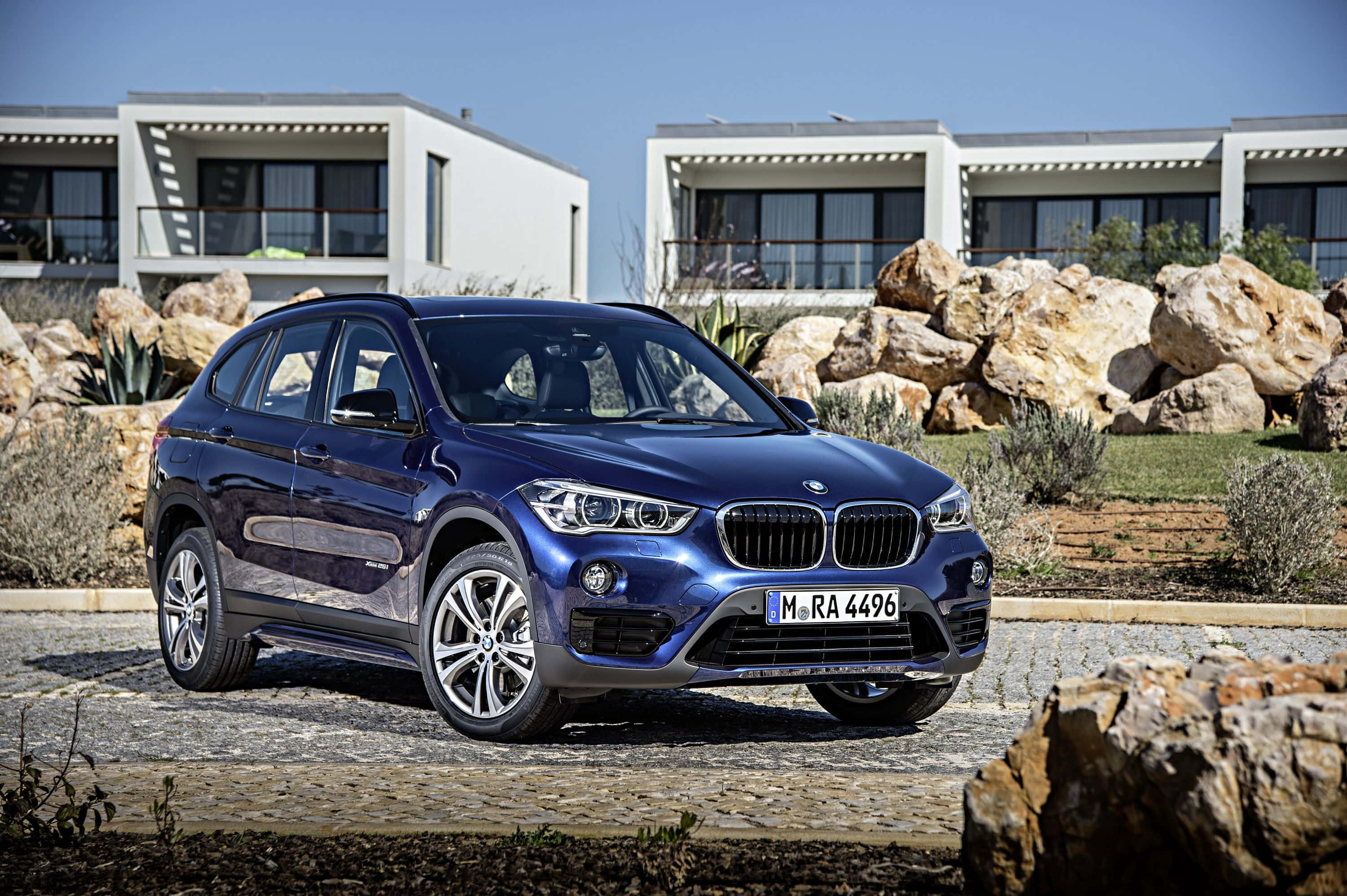 Bmw I8 桌布 壁紙 — 一架時尚的藍色BMW X1 SUV停泊在鵝卵石車道上，周圍有現代白色房屋和岩石沙漠景觀，天空清澈湛藍。