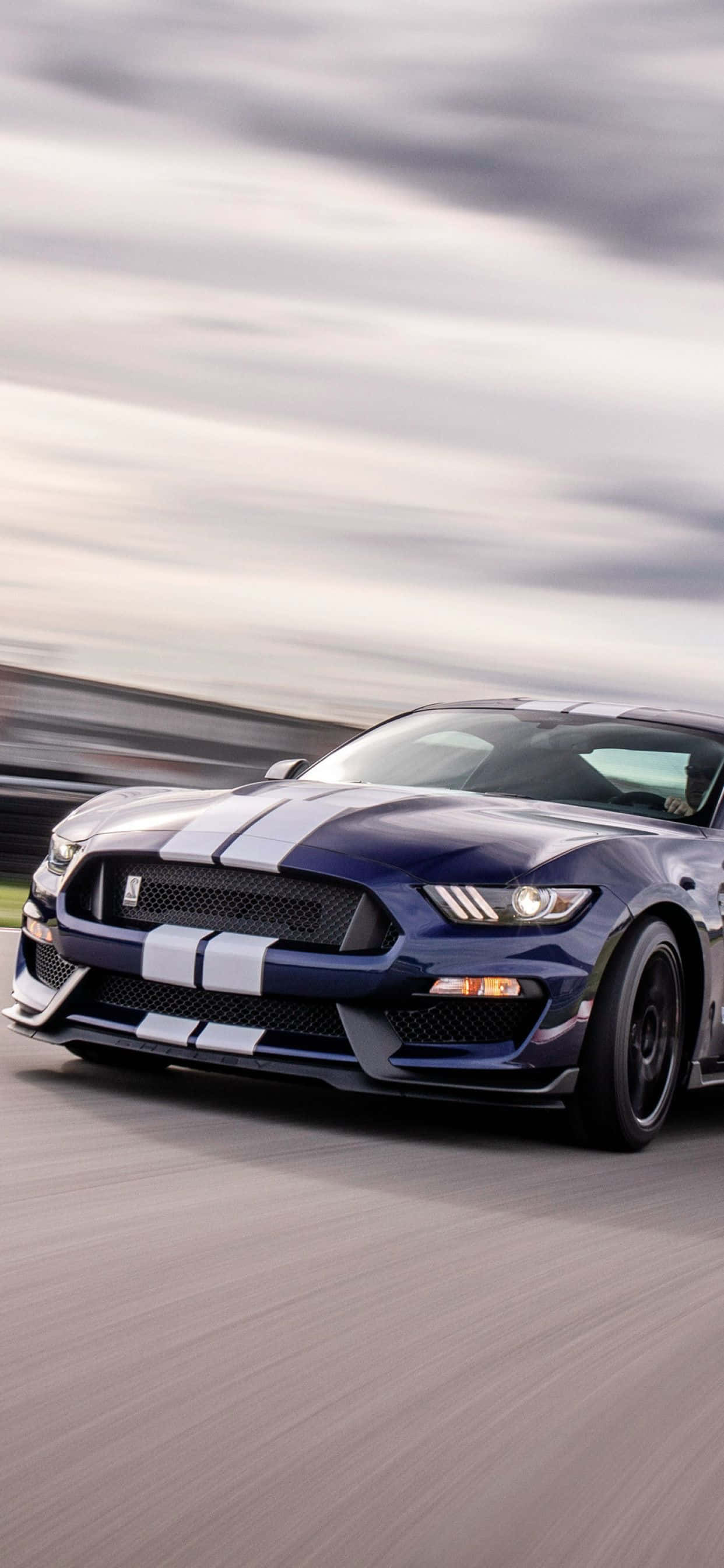 Ford Mustang Shelby Gt350 桌布 壁紙 — 一架時尚的藍色福特野馬，帶有白色賽車條紋，在陰天天空下沿著模糊的賽道飛馳。