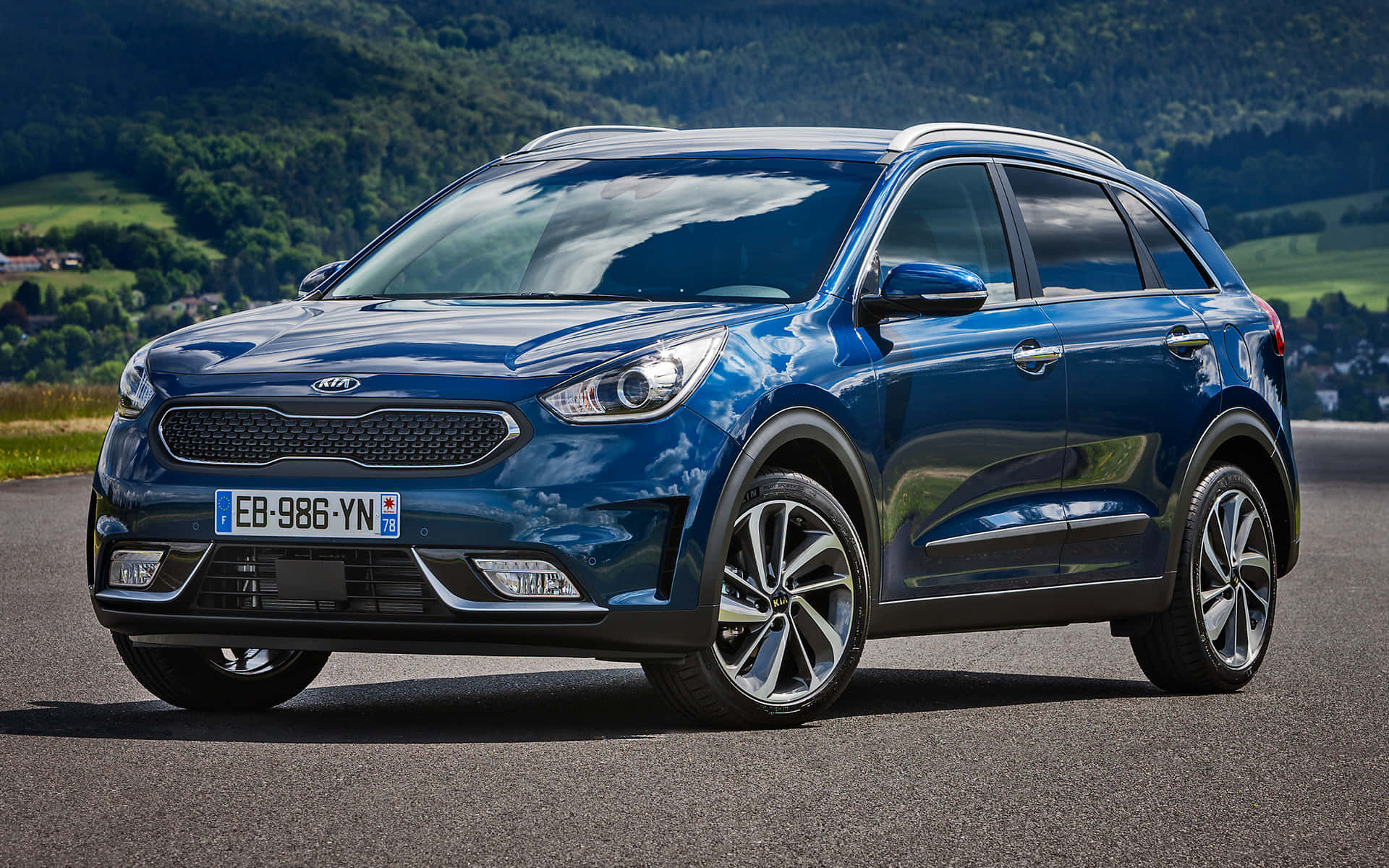 Kia Niro 桌布 壁紙 — 一輛時尚的藍色Kia SUV停在瀝青路上，背景是起伏的綠色山丘和樹木，在明亮的日光下。