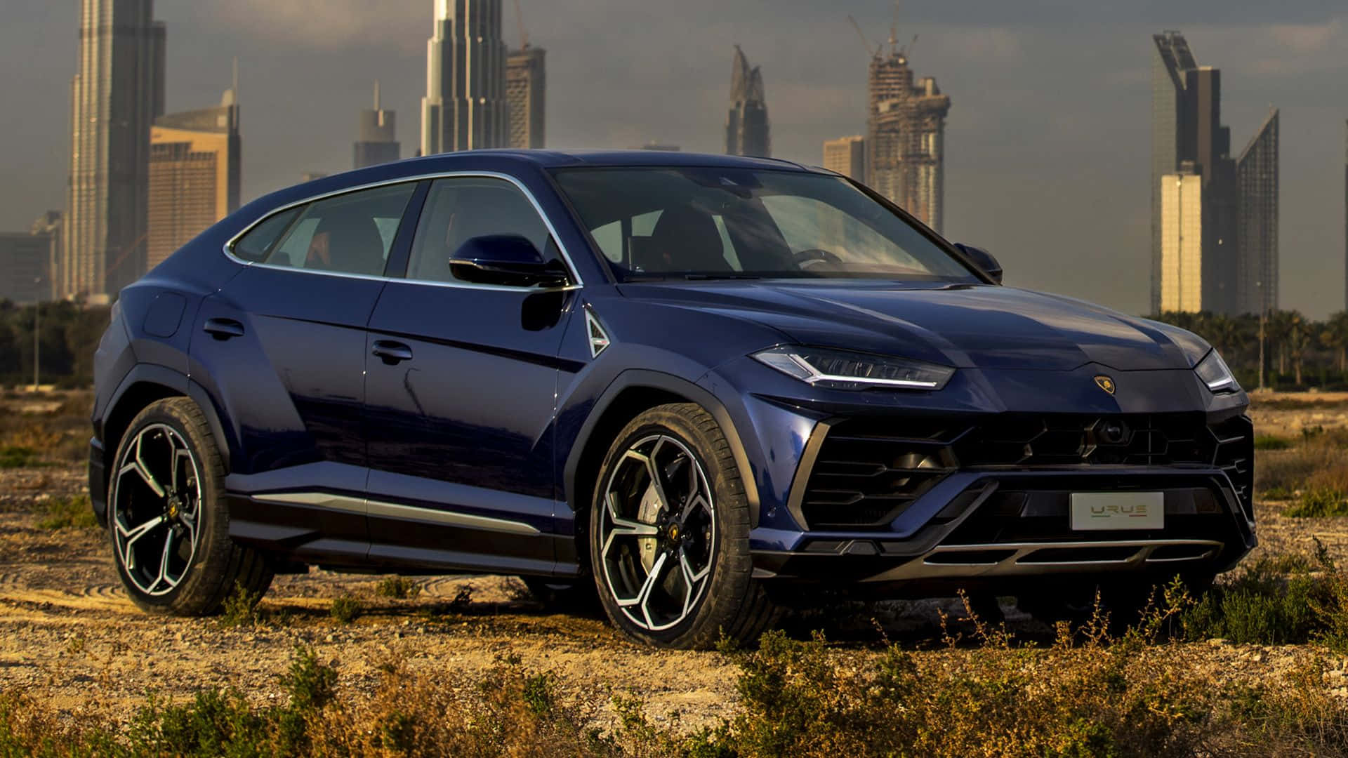 Lamborghini Urus 桌布 壁紙 — 一架時尚的藍色林寶堅尼 Urus SUV 立於乾草上，背景是現代城市天際線。