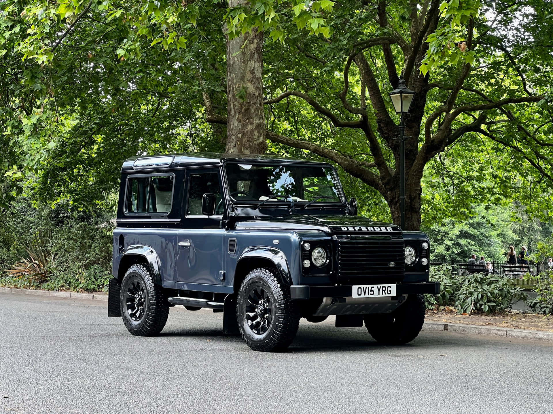 Land Rover Defender 桌布 壁紙 — 一輛時尚的藍色 Land Rover Defender 停泊在陽光普照、綠樹成蔭的公園道路上。