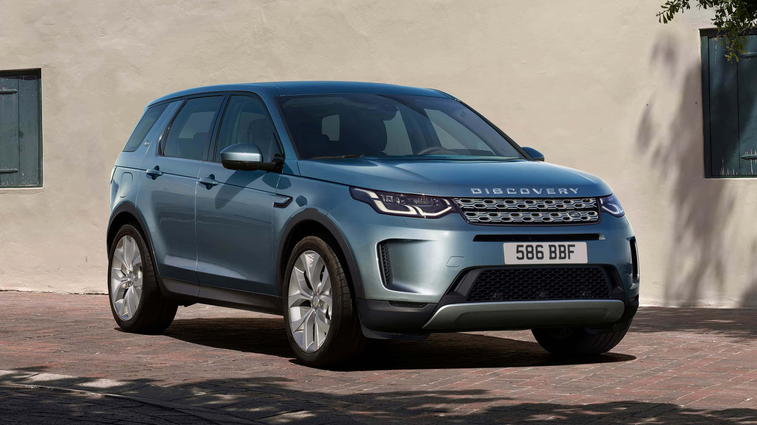 Land Rover Discovery 桌布 壁紙 — 一輛時尚的藍色 Land Rover Discovery 停泊在陽光明媚的磚砌車道上，旁邊是帶綠色百葉窗的米色灰泥牆。