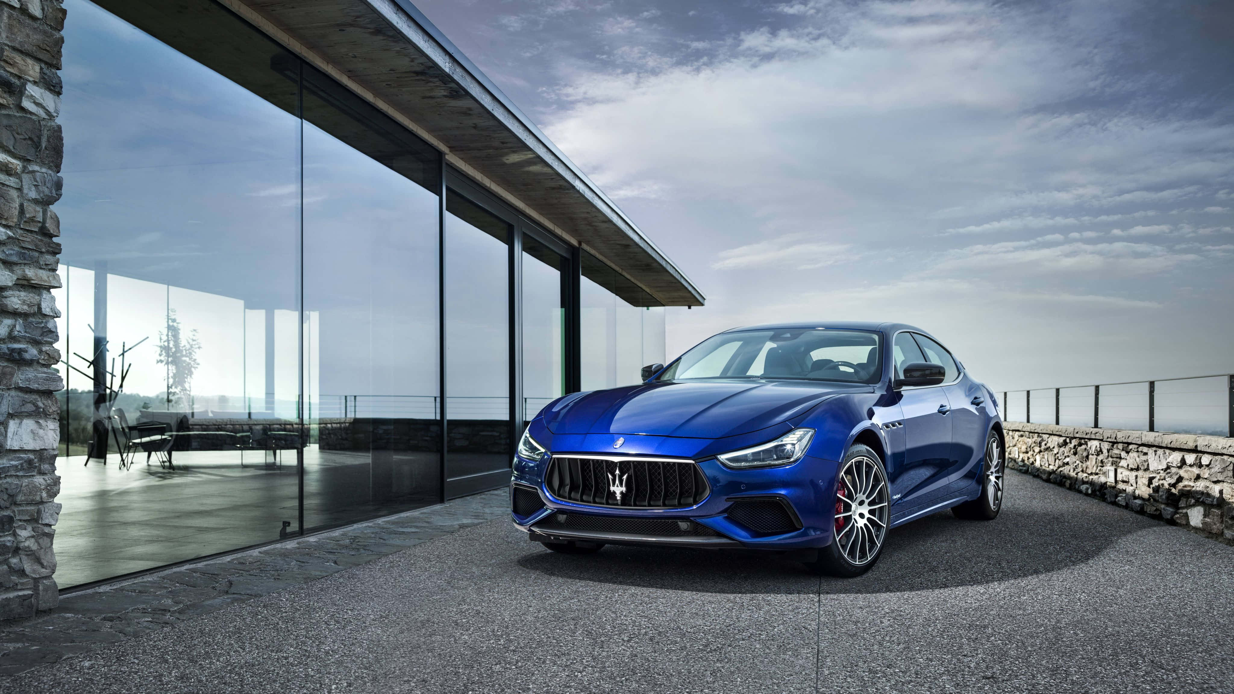 Maserati Ghibli 桌布 壁紙 — 一架時尚的藍色瑪莎拉蒂跑車停泊在石頭車道上，旁邊是一座現代玻璃屋，天空多雲。
