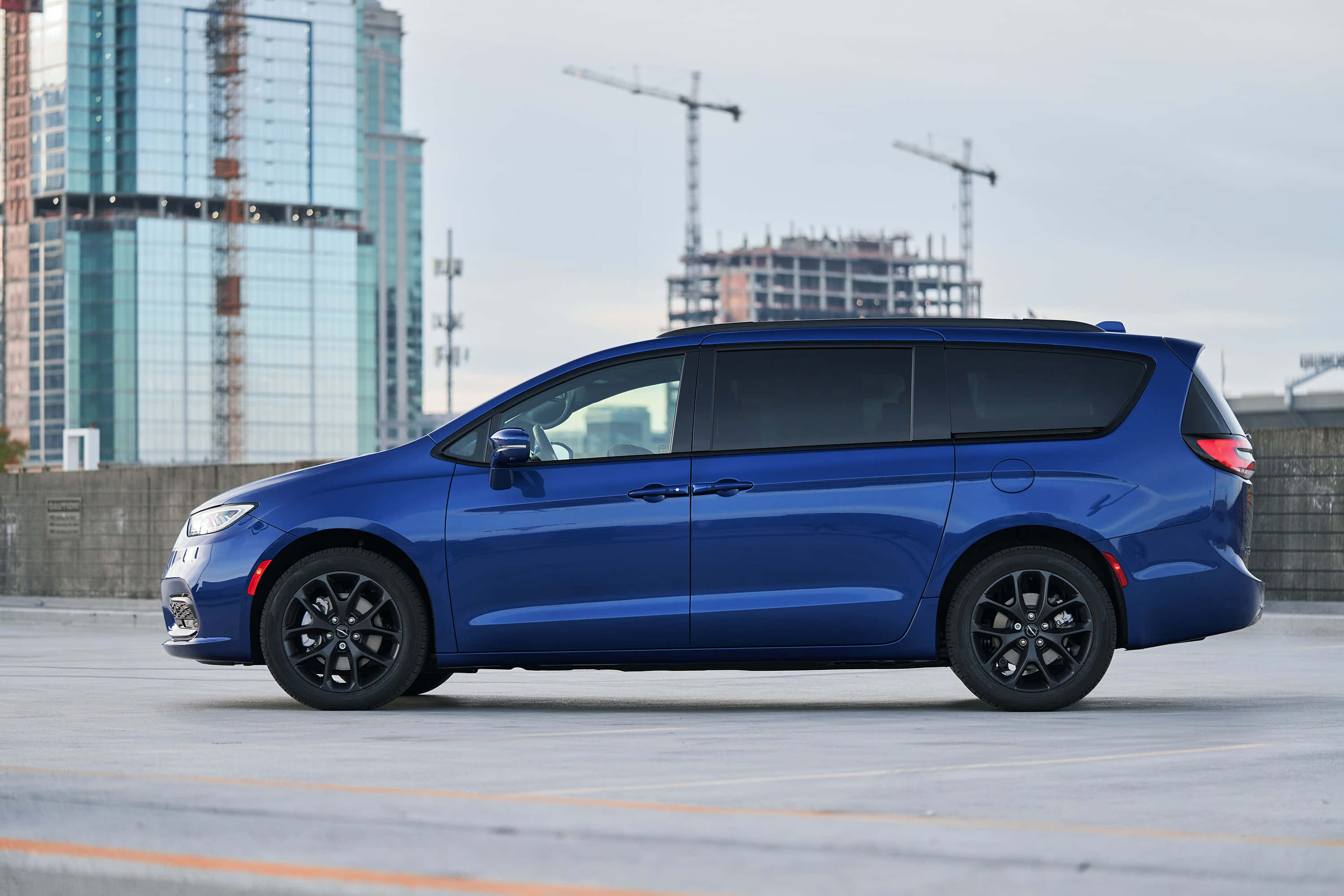 Chrysler Pacifica 桌布 壁紙 — 一架時尚的藍色小型貨車配黑色輪圈，停泊在城市空地，背景是玻璃摩天大樓和建築起重機。