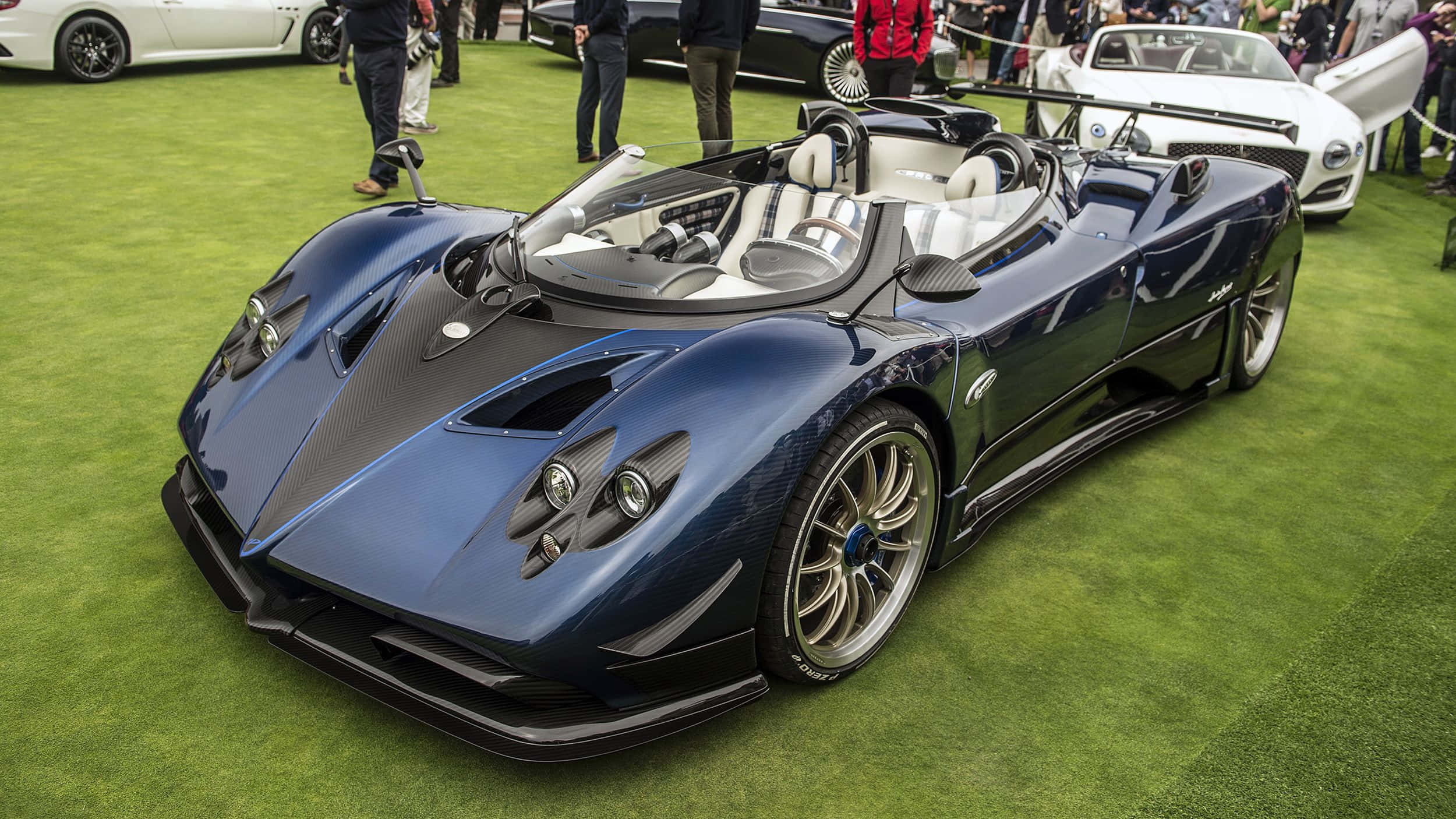 Pagani Zonda Hp Barchetta 桌布 壁紙 — 一輛流線型的藍色帕加尼 Huayra 開篷跑車在車展的綠色地毯上閃耀著明亮燈光，人們正在欣賞它。