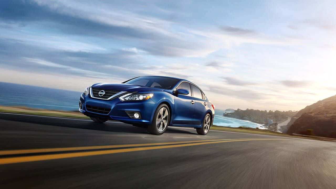 Nissan Altima 桌布 壁紙 — 一輛時尚的藍色轎車沿著海岸公路飛馳，海浪拍打懸崖，天空呈現柔和的金色日落。