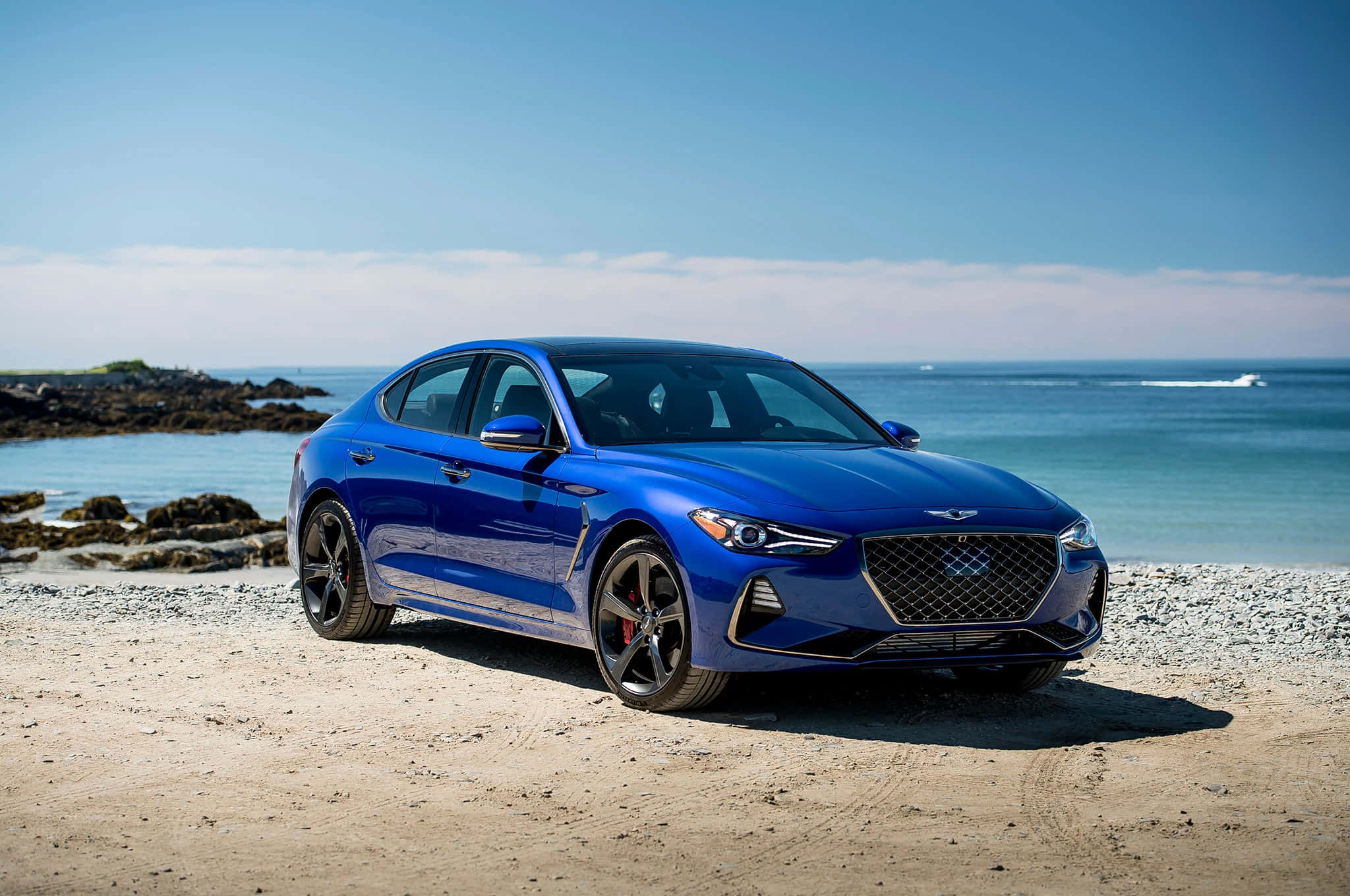 Genesis G70 桌布 壁紙 — 一架時尚的藍色跑車停泊在陽光明媚的海灘上，背景是海洋和岩石海岸。
